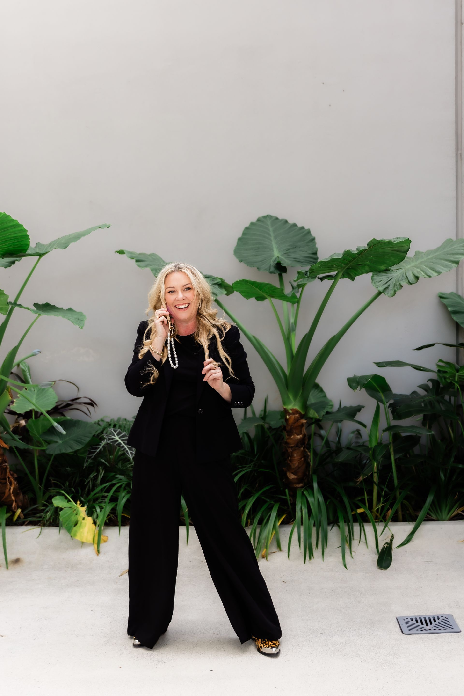 Woman in black suit smiles while on phone in front of green plants and a concrete wall.