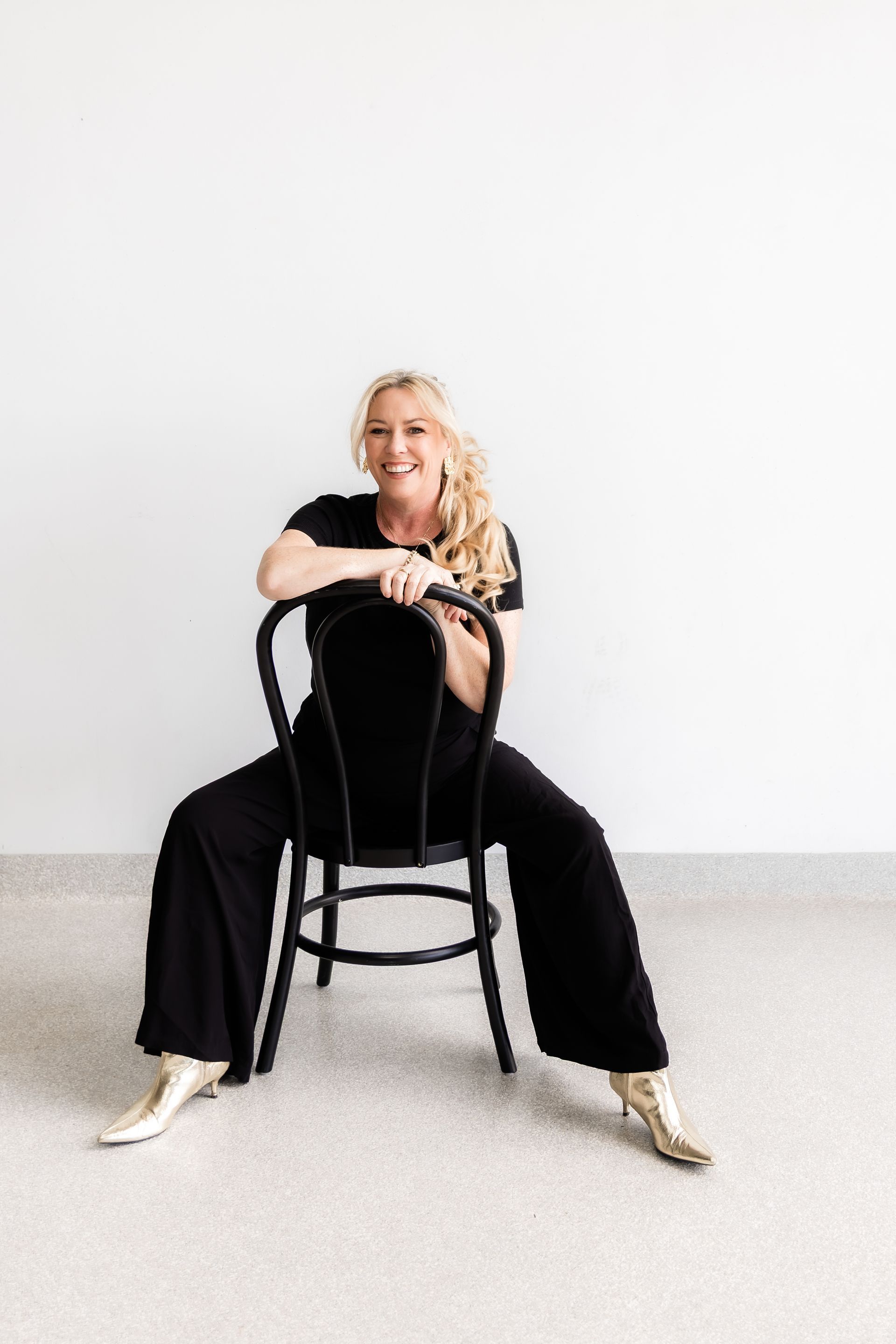 Woman seated on a black chair, arms resting on back. Wearing black outfit, gold boots, and smiling. White background.