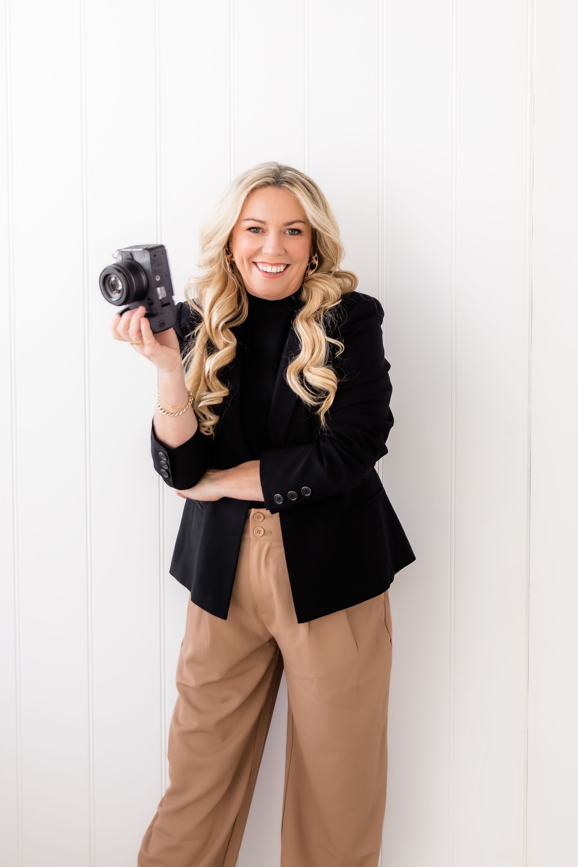 Woman with camera smiles, wearing black blazer, tan pants, standing against white wall.