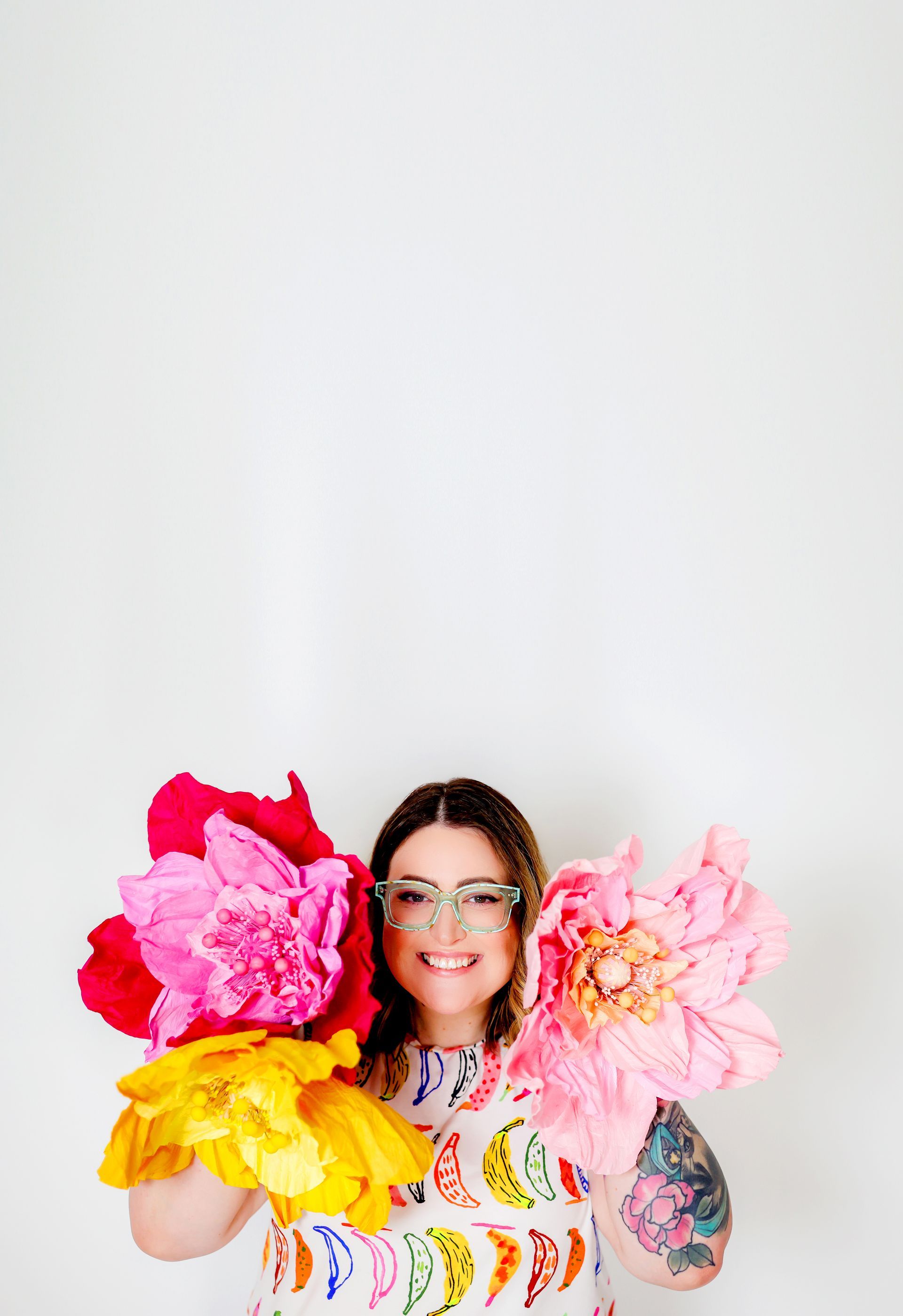 Woman holding large paper flowers, smiling, wearing glasses and patterned dress. Against a white background.