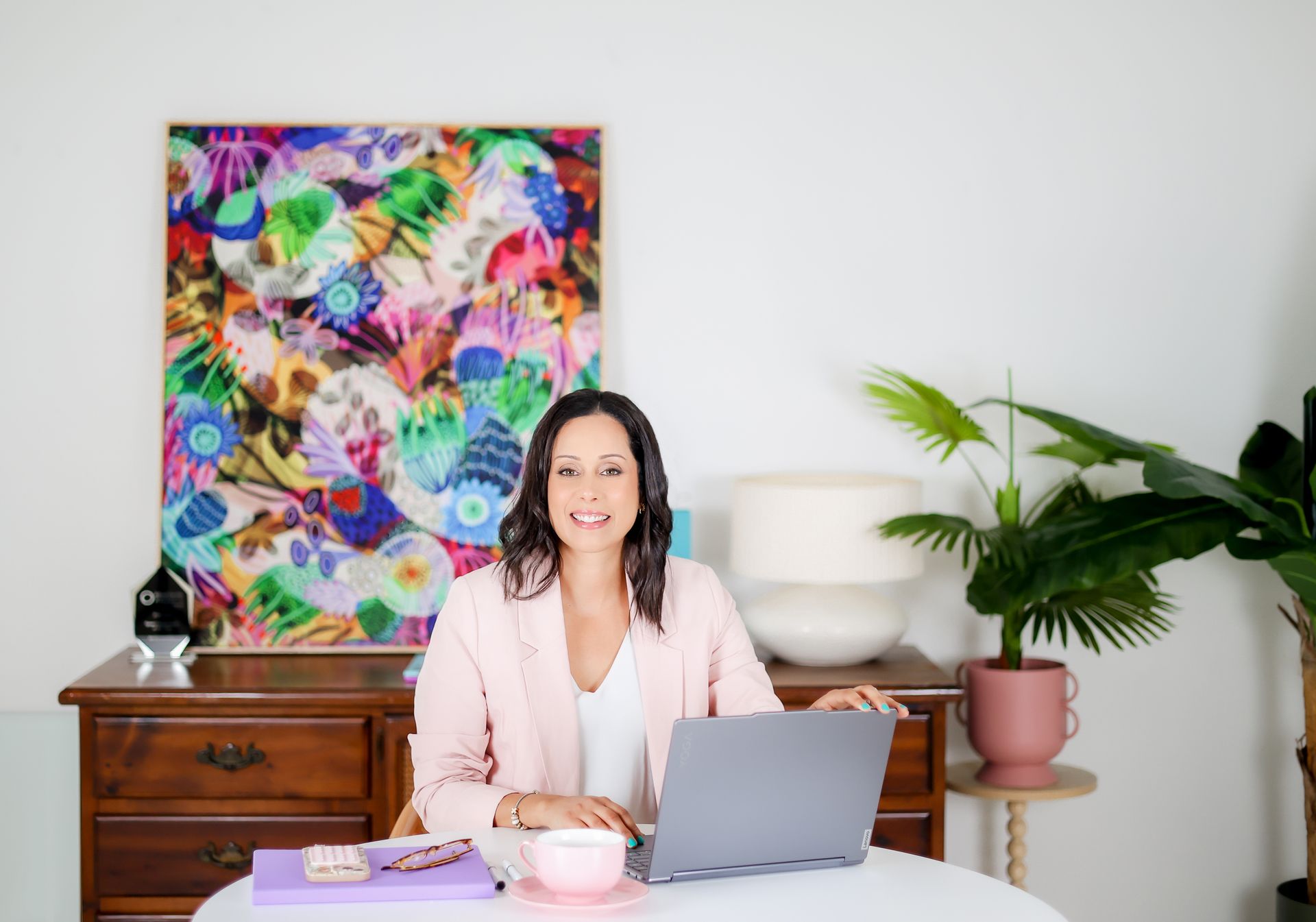 Woman in a pink blazer smiles at the camera while working on a laptop at a white table.