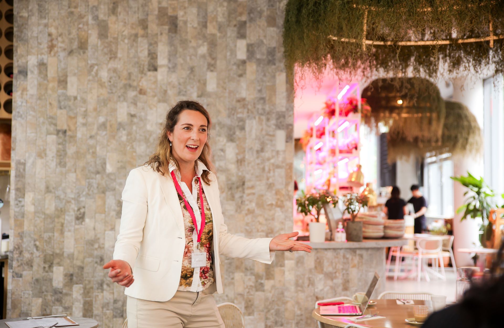 Woman in cream blazer speaking with hands open; neutral-toned wall and hanging plants in background.