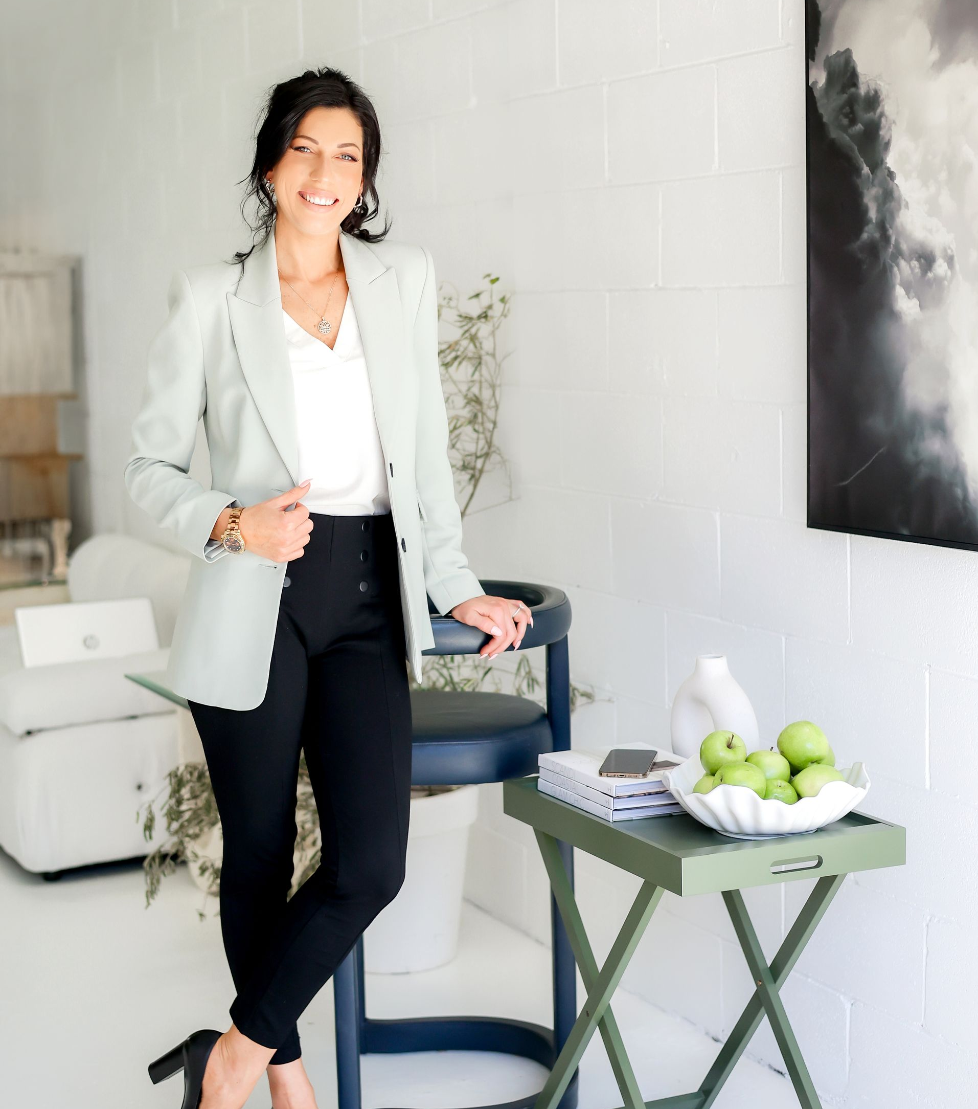 Woman in light green blazer, black pants, smiles next to a small green table with apples, a chair, and a painting.