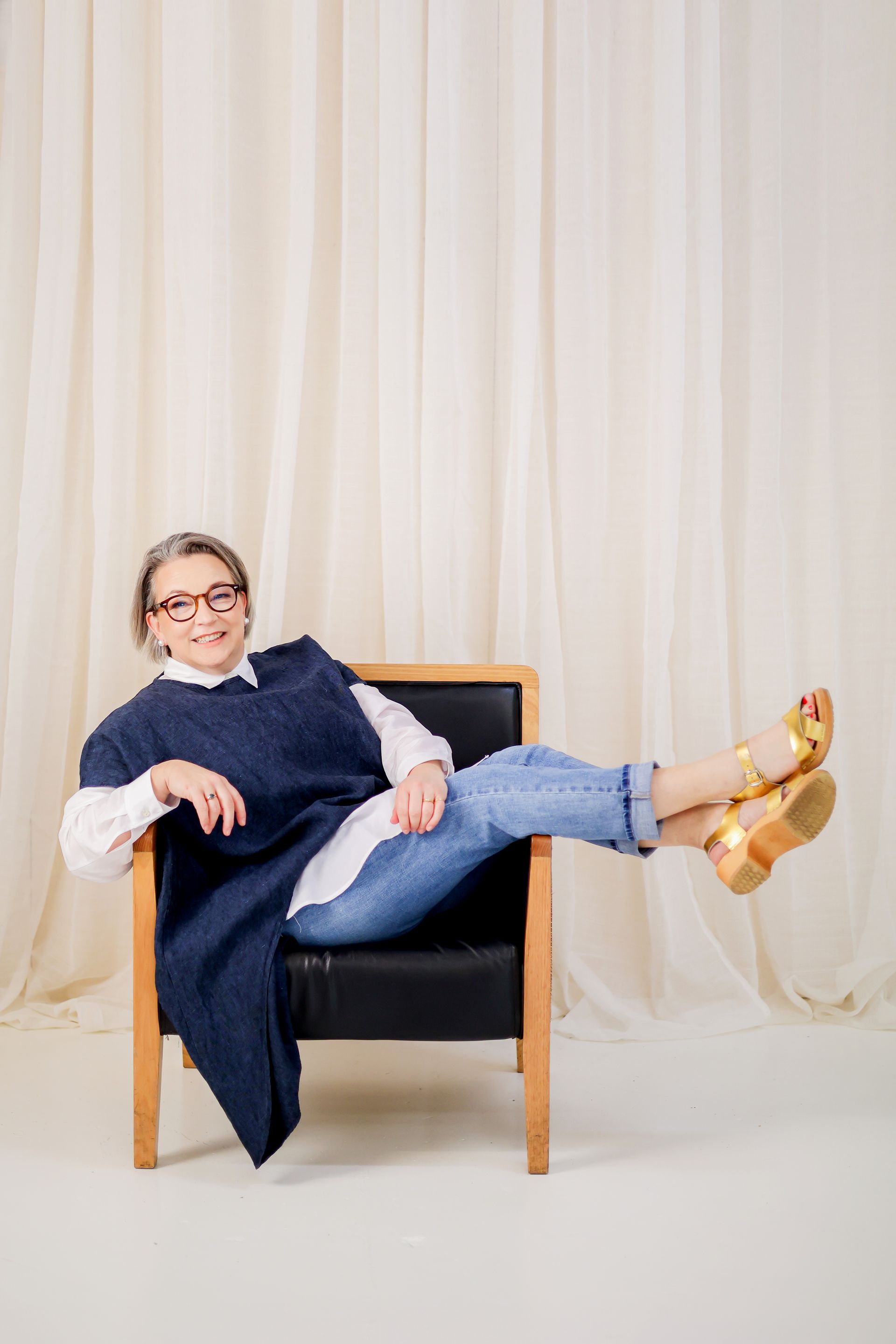 Woman in blue top, jeans, and gold sandals, seated in a chair, legs up, smiling. Beige curtain background.