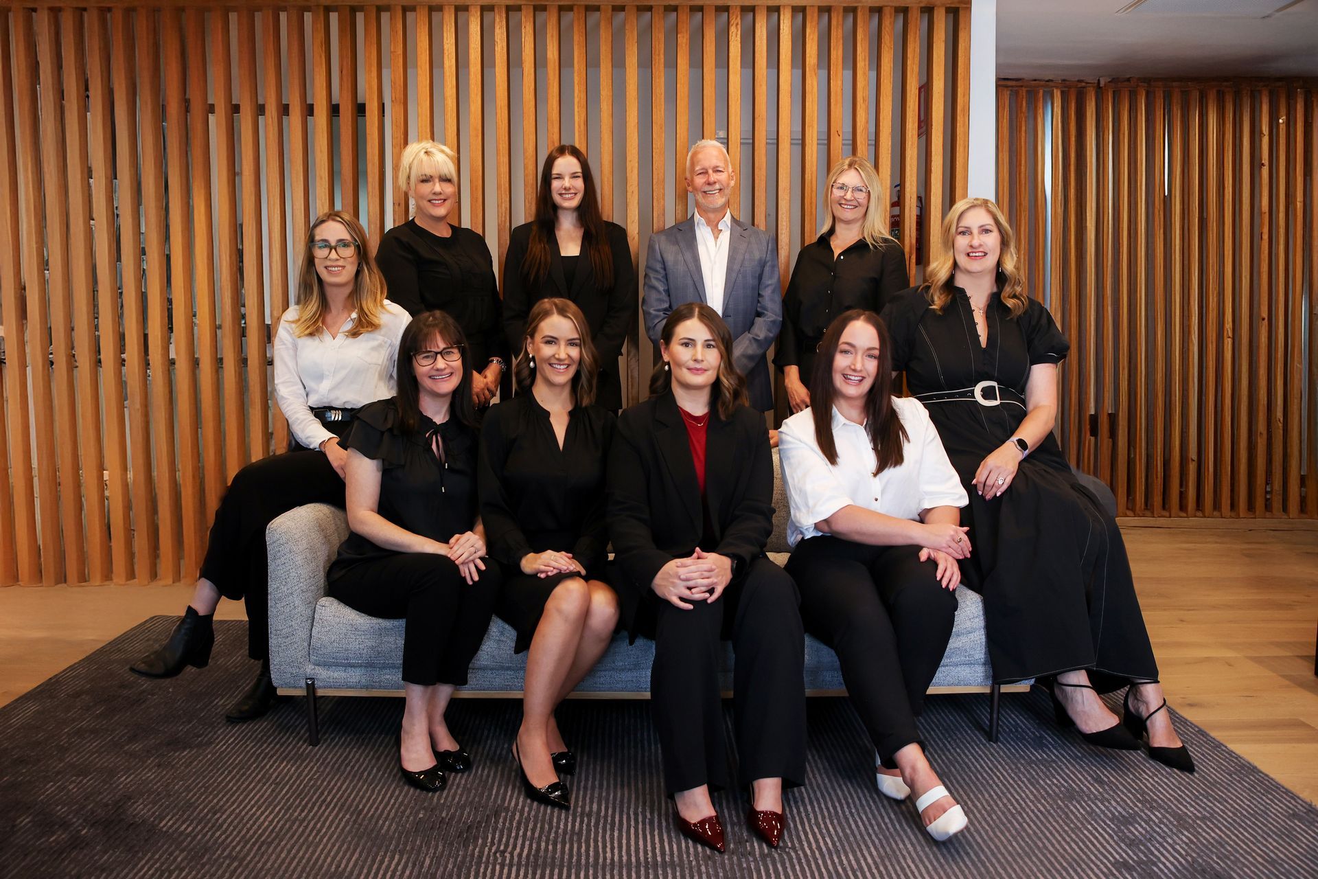 Group of 11 people posing for a photo. They are seated/standing in front of a wood-paneled wall.