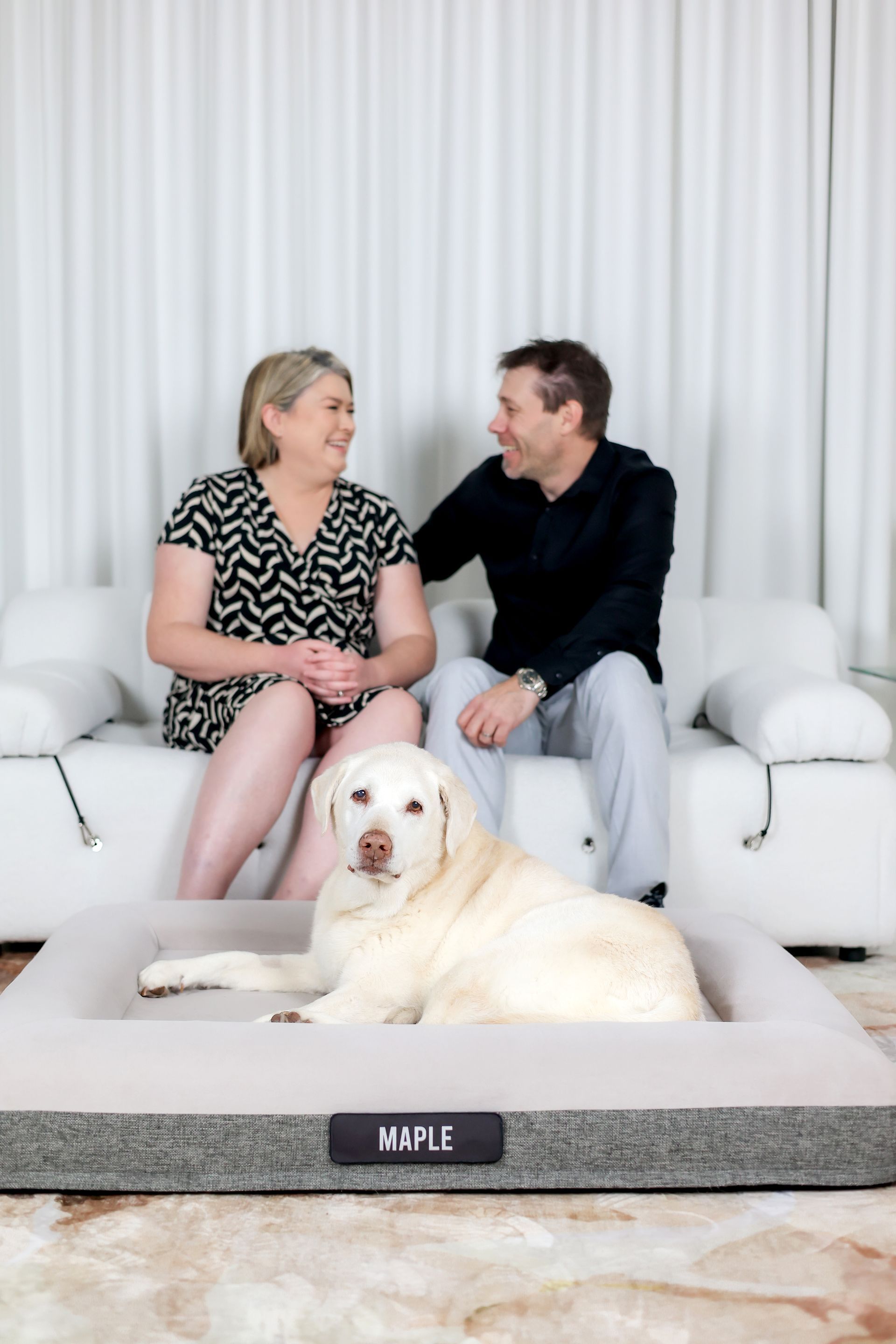 Couple on sofa, smiling at each other. Labrador dog in bed in front. Neutral-toned room, soft light.