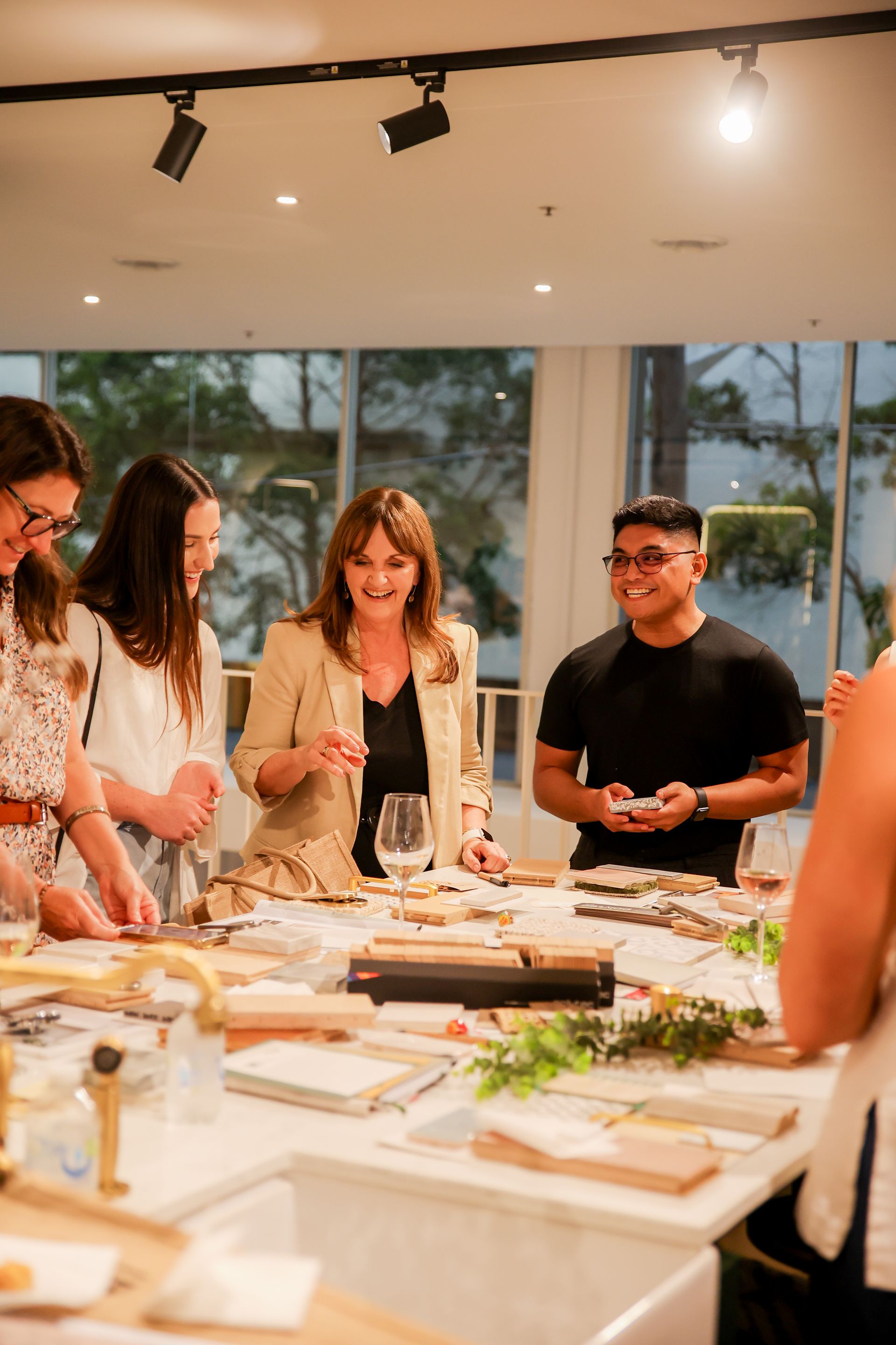 People gather around a table, smiling and interacting, in a well-lit workshop setting.