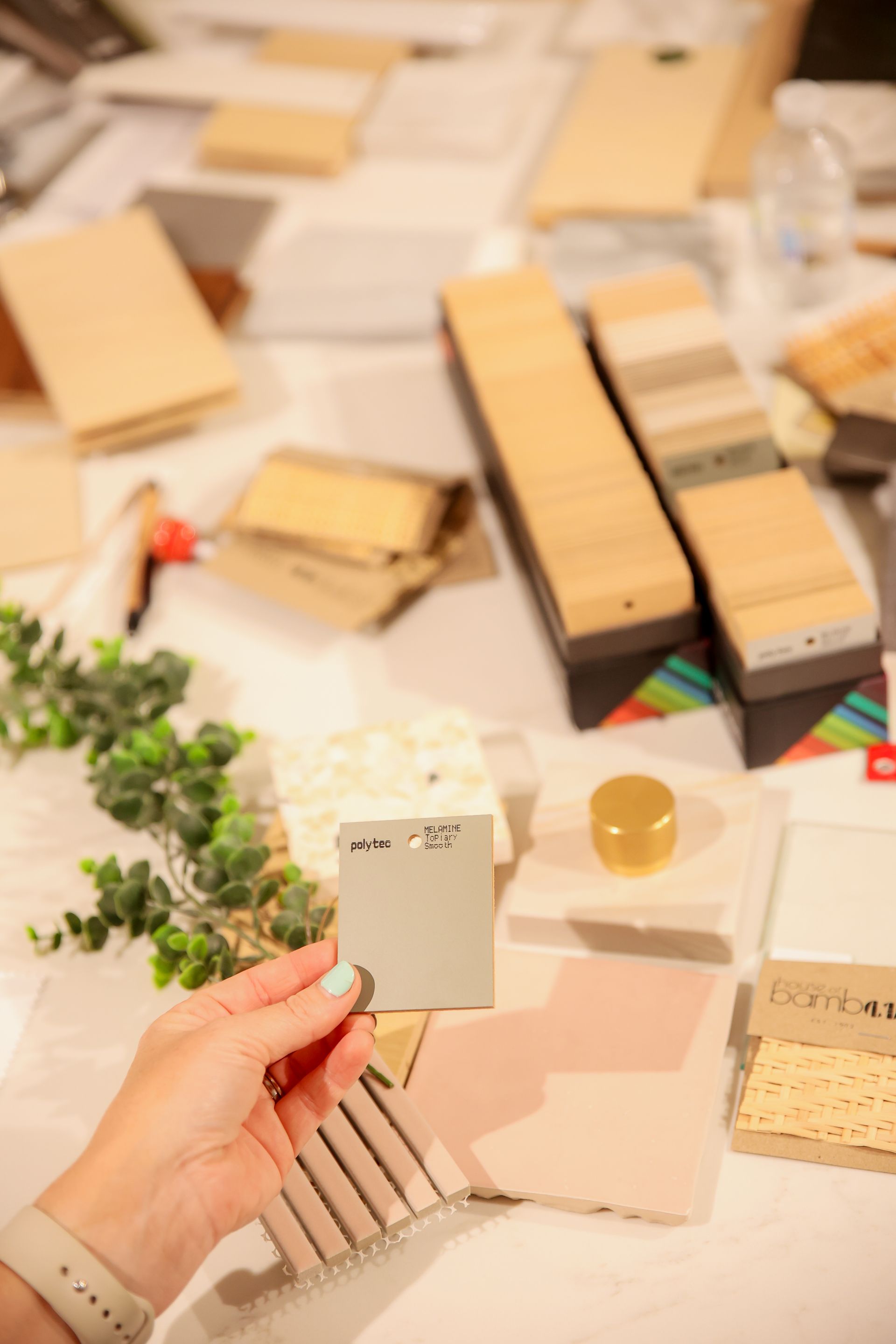 Hand holding a gray paint chip over pink and textured samples on a table with other design materials.