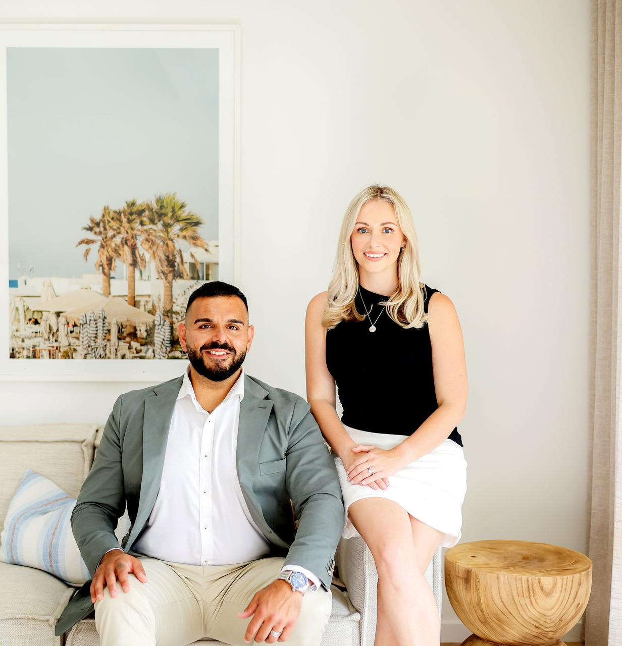Man and woman sitting, smiling. Man in blazer, white shirt, khaki pants. Woman in black top, white skirt. Beach scene art.