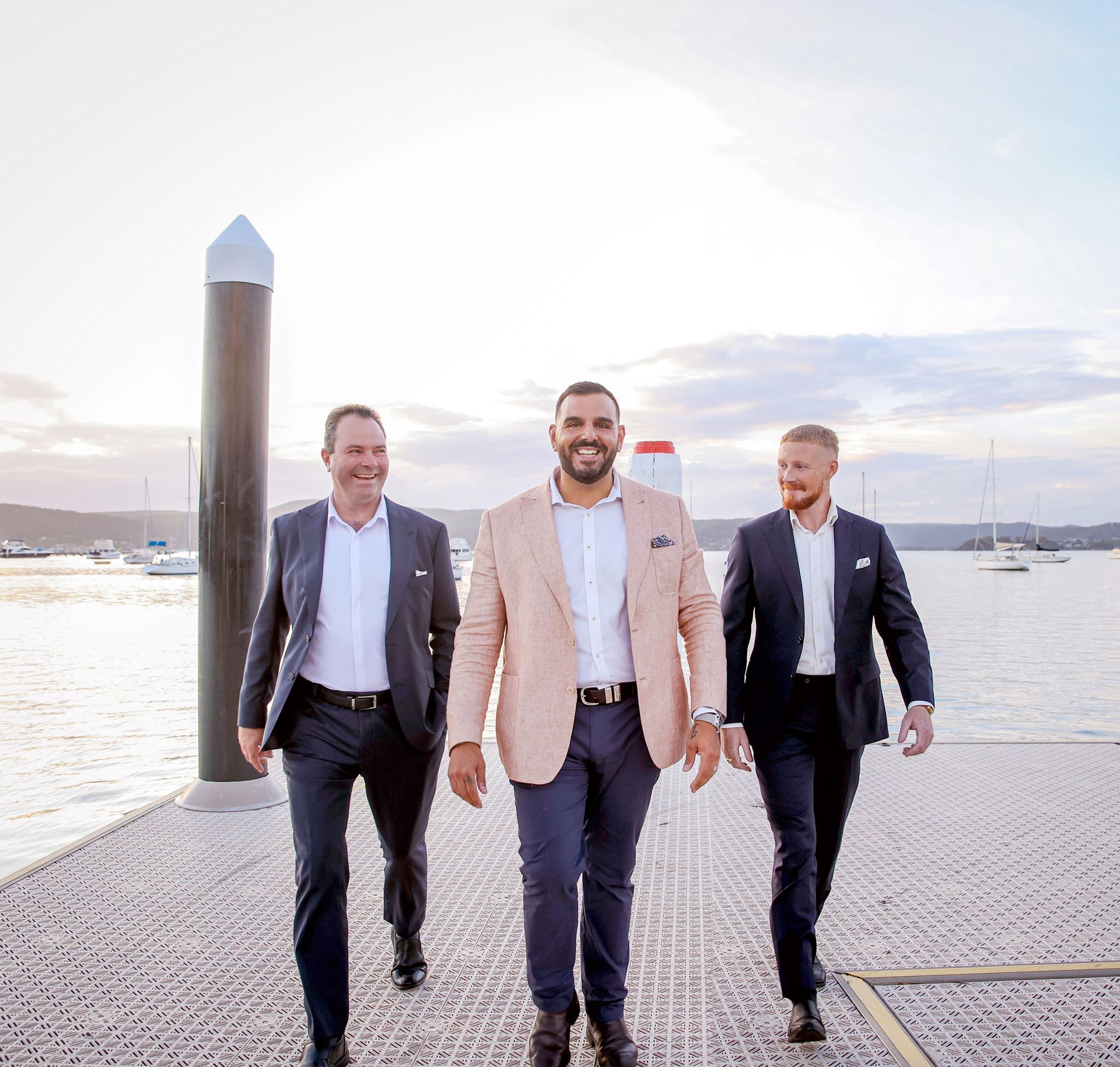 Three men in suits walking on a dock with a waterfront view.