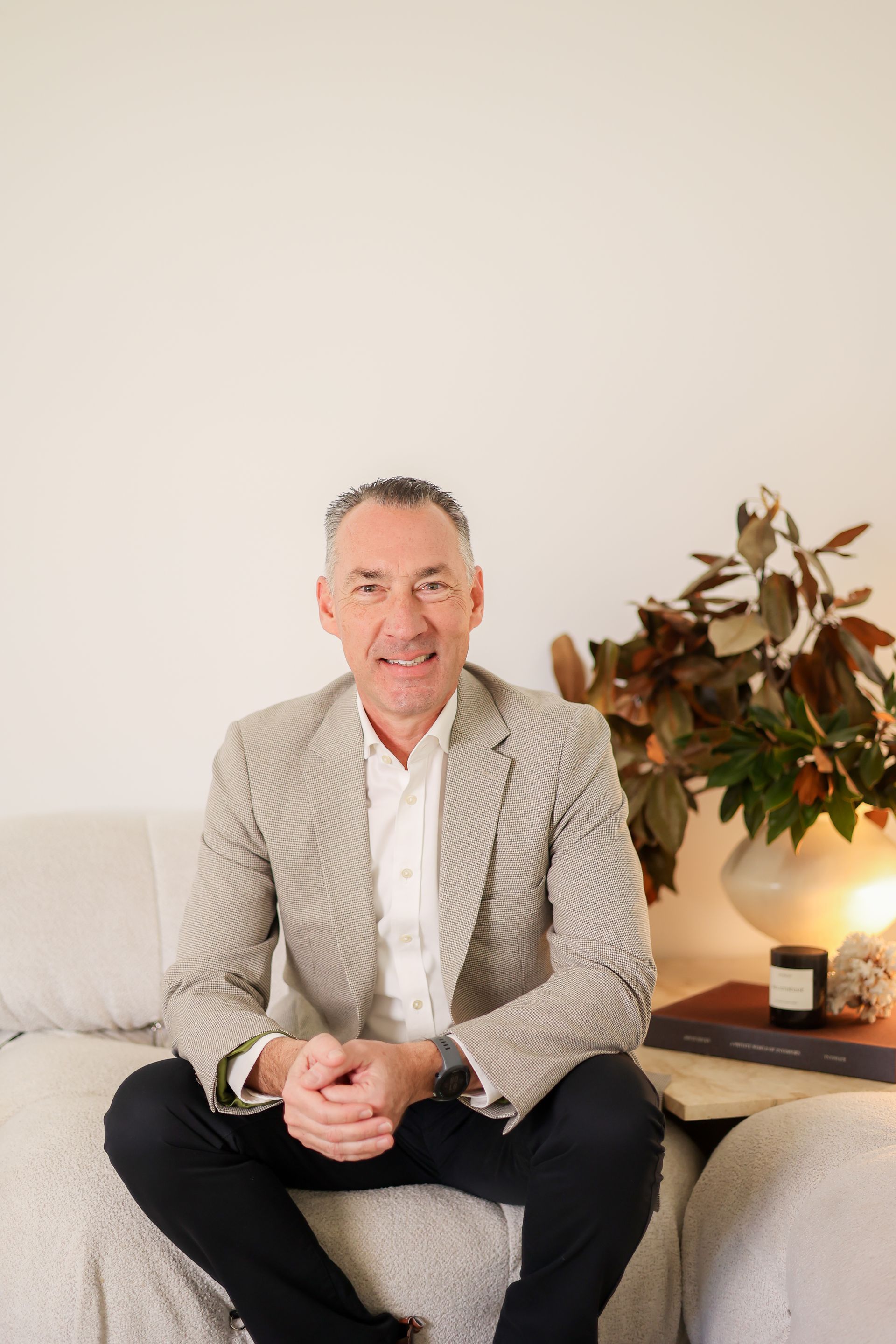 Man in a patterned jacket sits on a cream couch, smiling. Next to him is a table with a floral arrangement.
