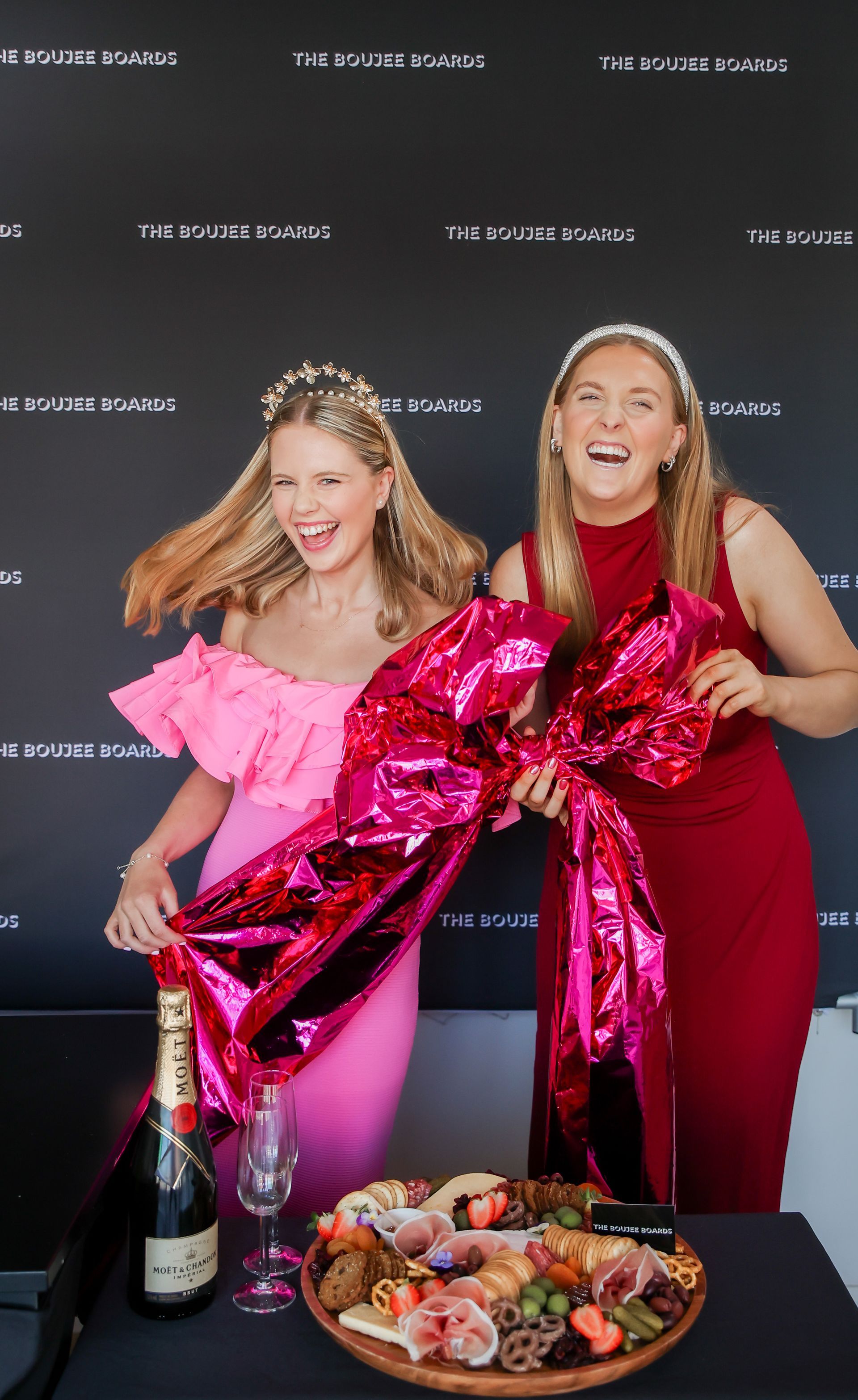 Two women smiling, holding a large pink bow, charcuterie board, champagne, black backdrop.