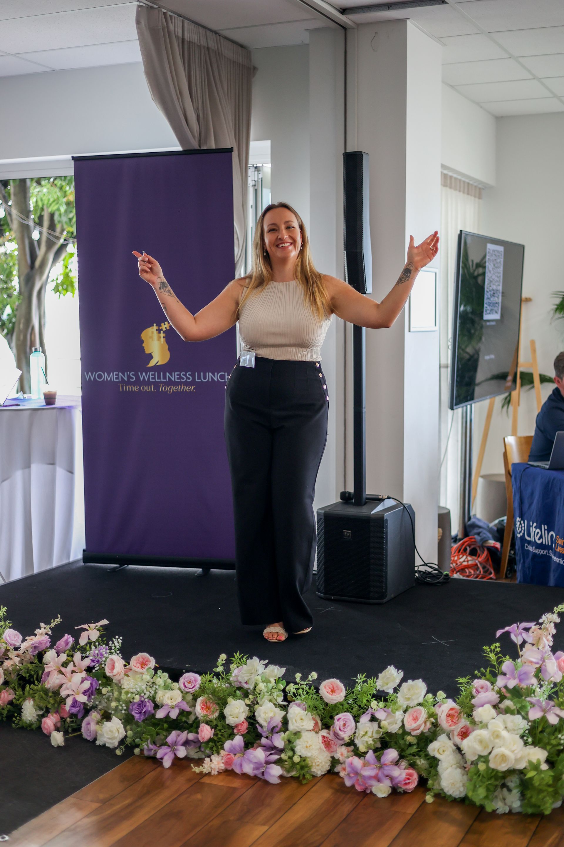 Woman giving a presentation on stage, arms outstretched, smiling. Purple banner, floral arrangement.