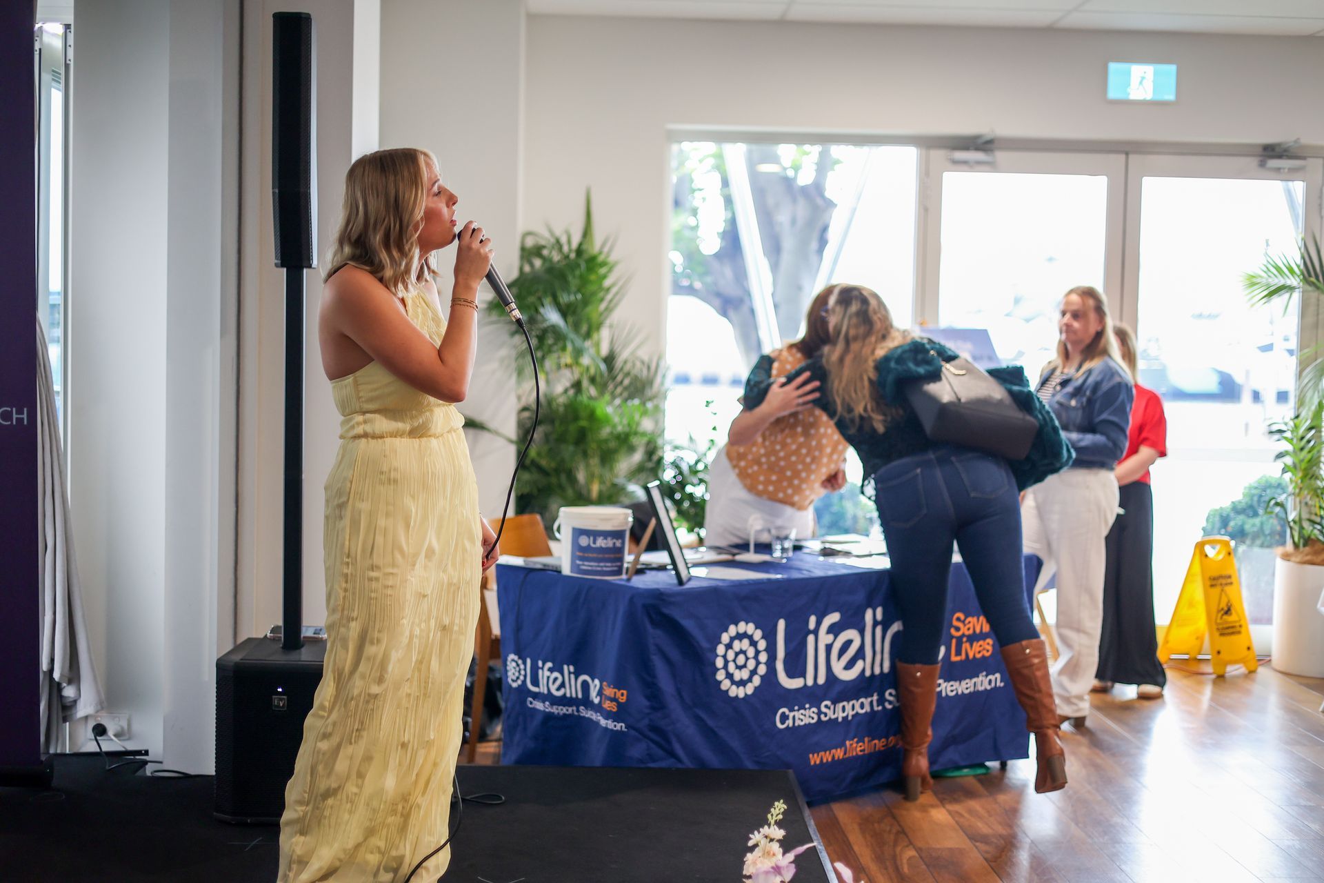 Woman singing at an event with a LifeLine table. Other people are in the background.