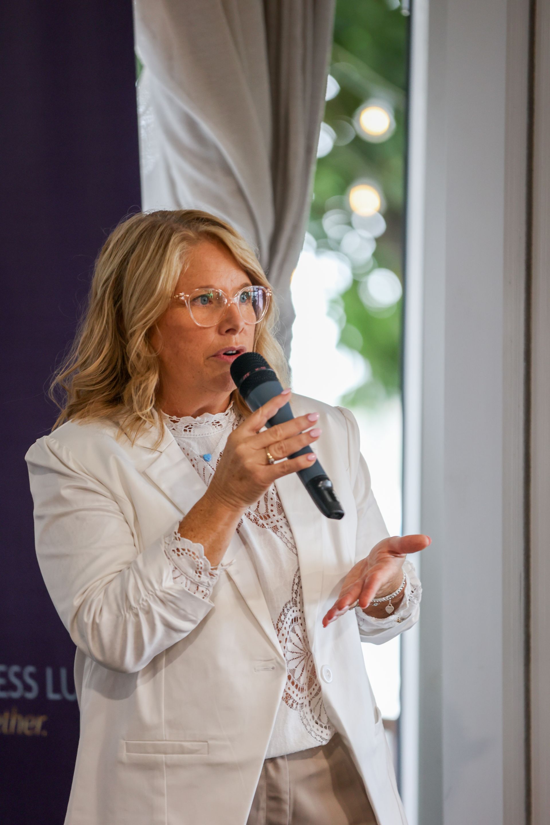 Woman in white blazer speaks into a microphone, gesturing with one hand; indoor event setting.