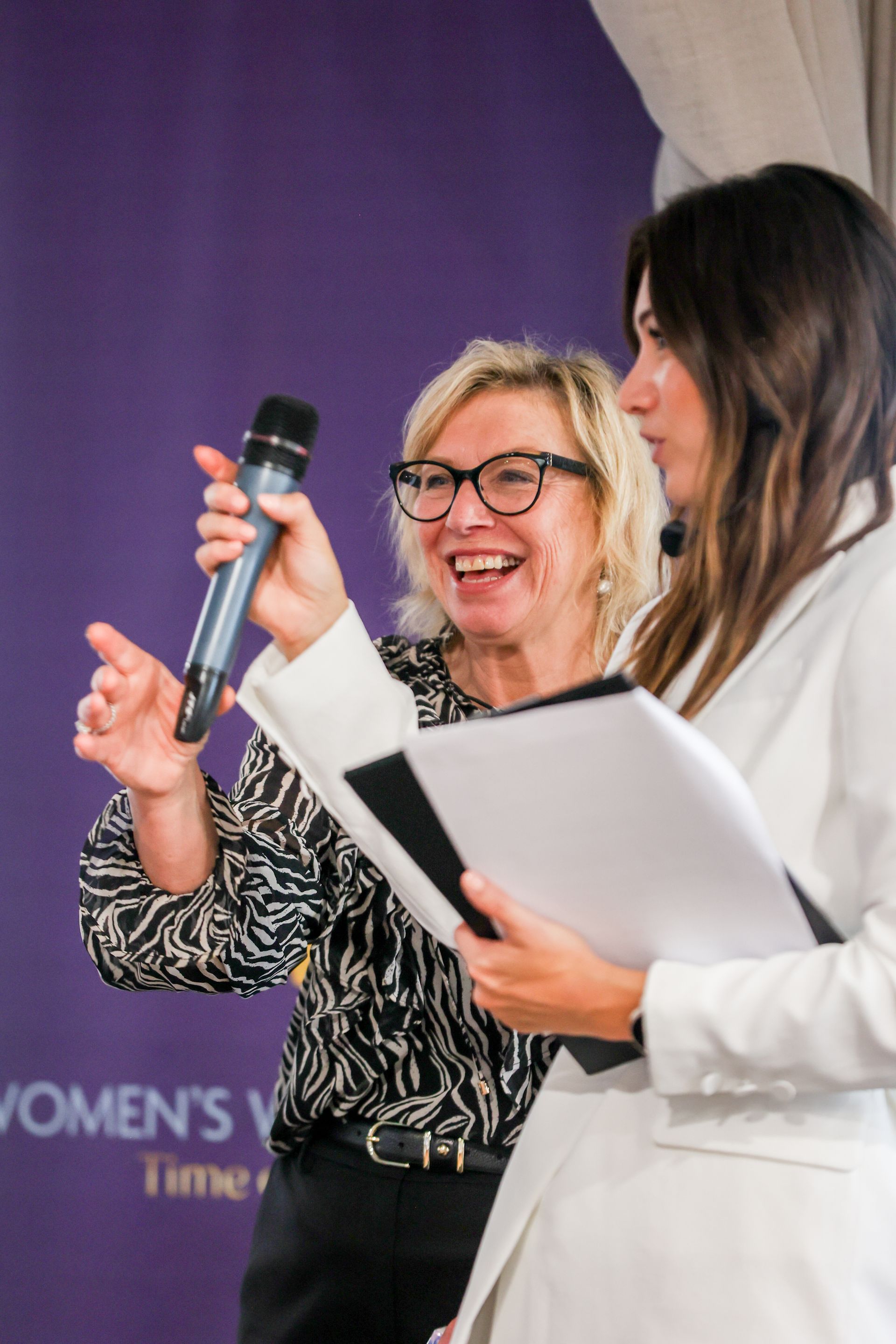 Woman with microphone, laughing, gestures at the right, another woman holds papers, purple backdrop.