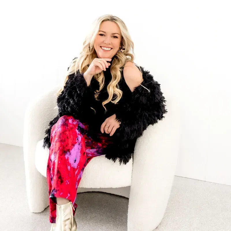 Woman in colourful pants and a black fringed jacket, smiling, seated in a white chair.