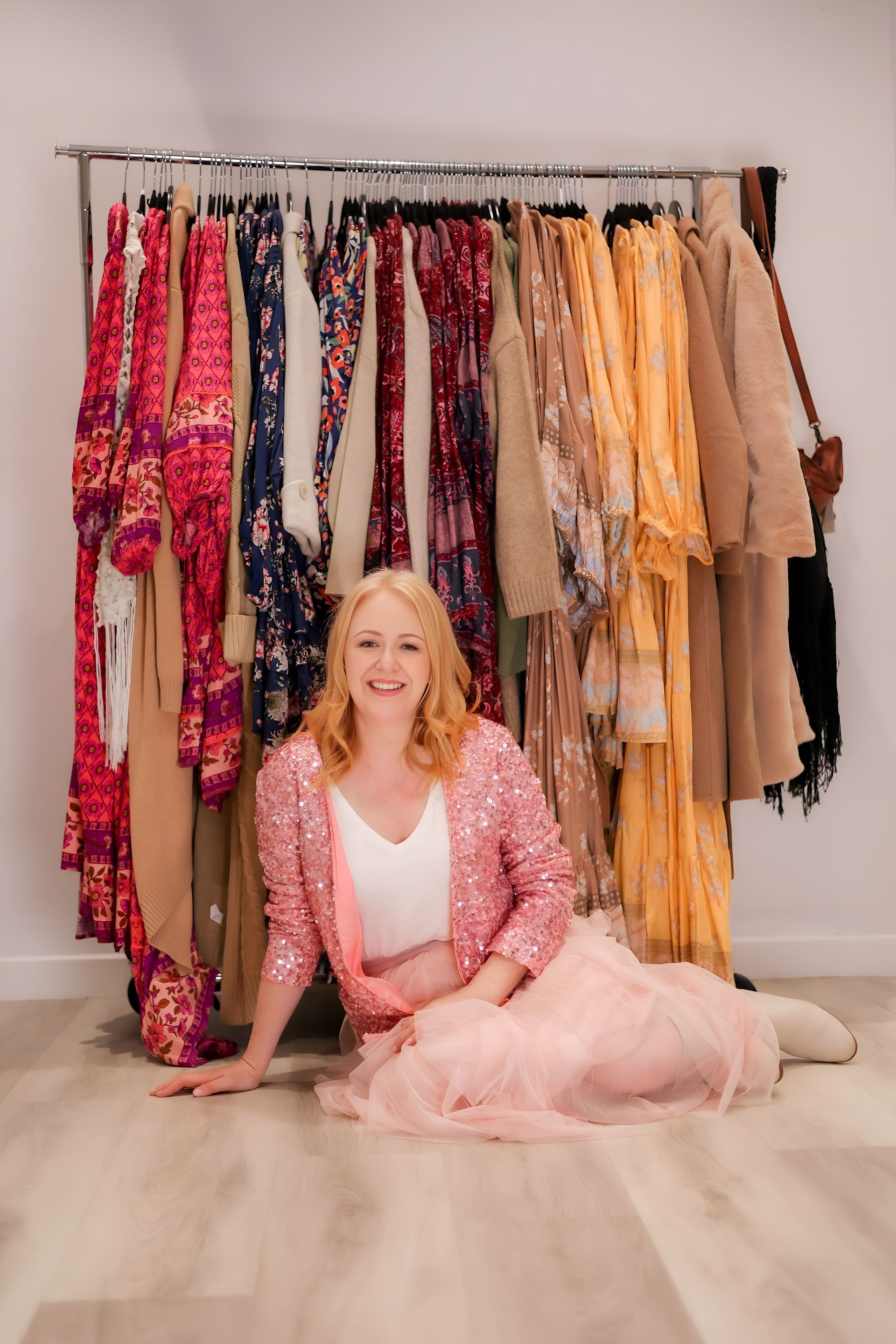 Woman in pink outfit sits in front of clothing rack. Smiling, blonde hair, colorful dresses.