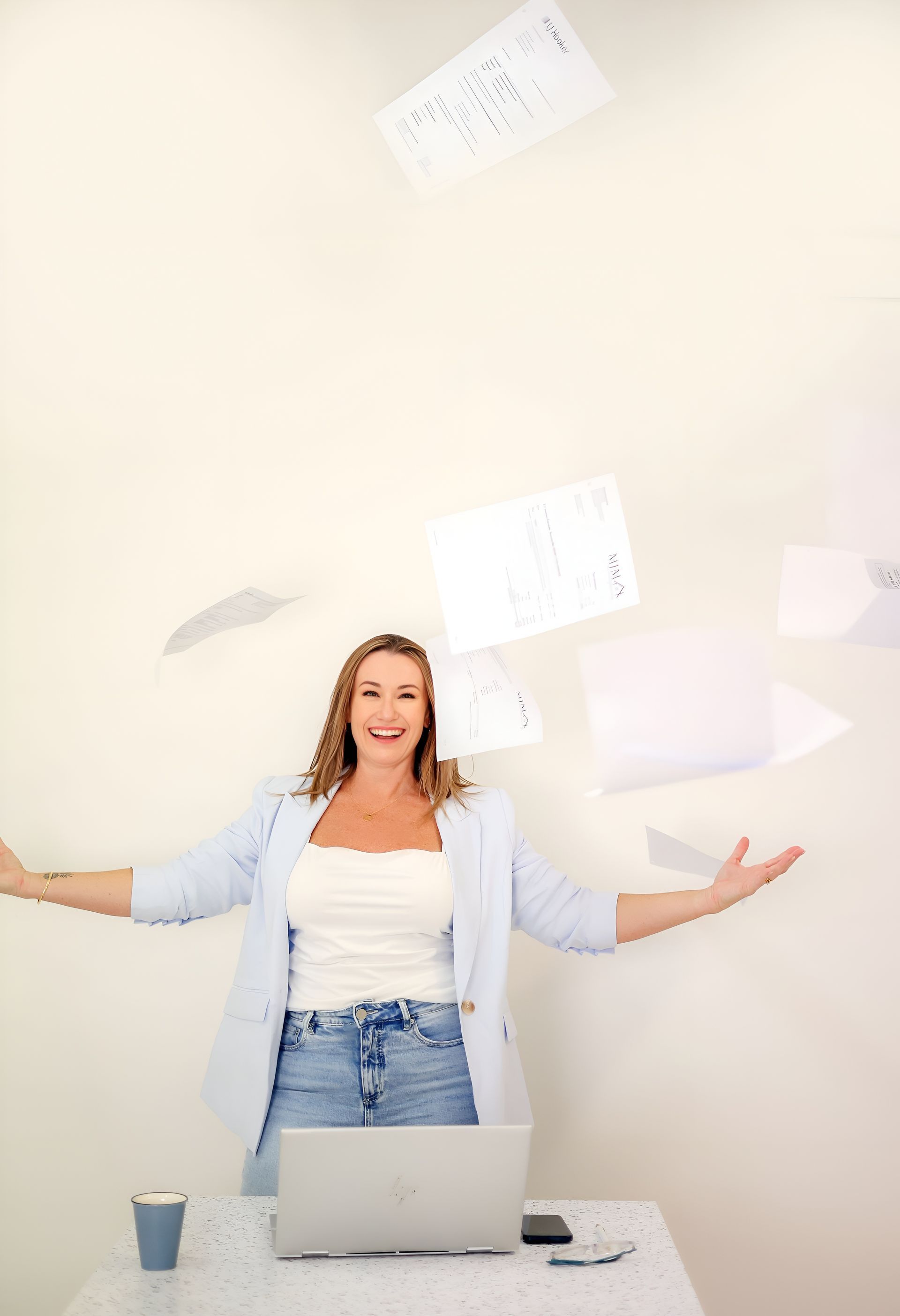 Woman tossing papers in the air with arms outstretched; laptop and desk in front of her.