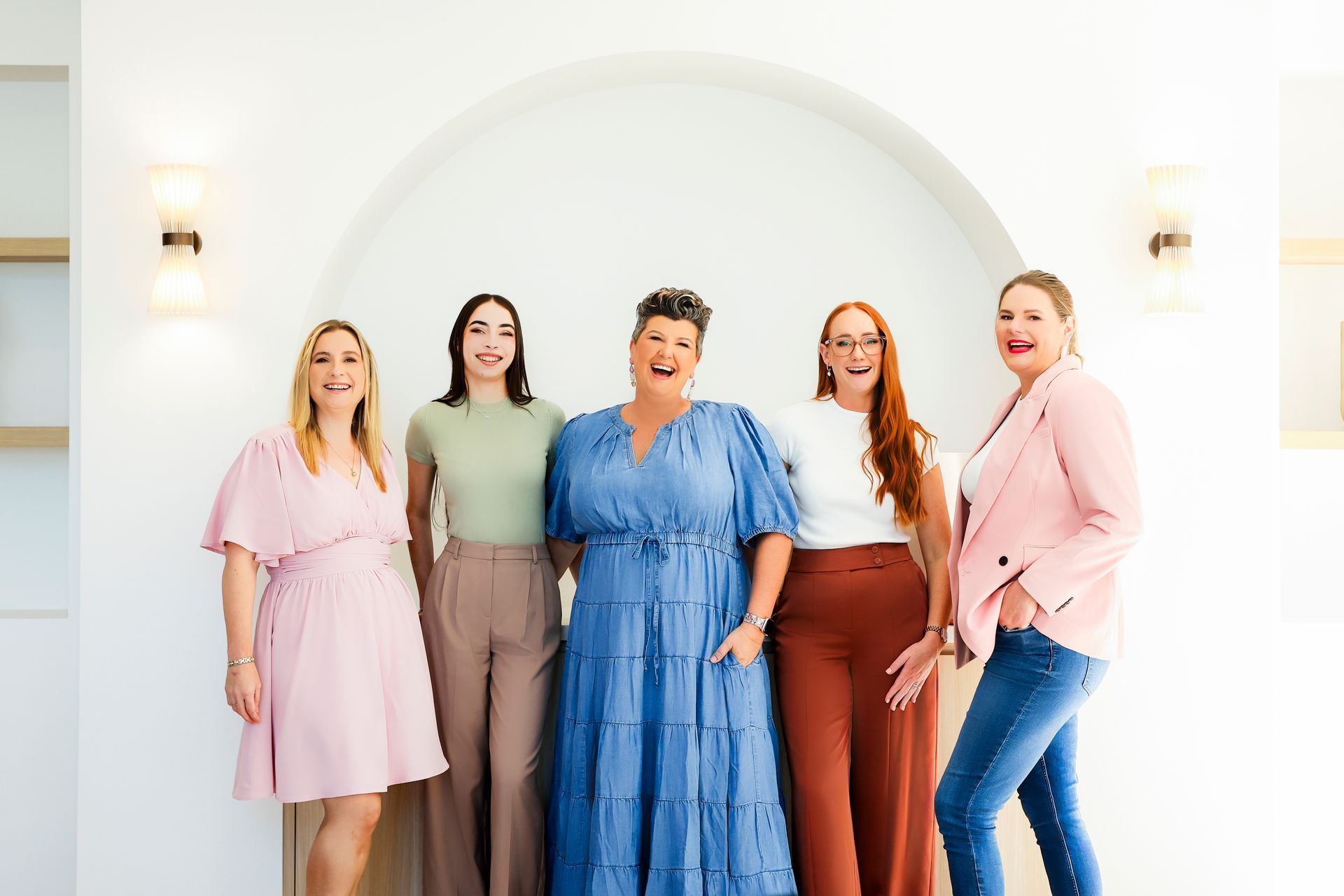 Five women smiling in a white archway, wearing various outfits.