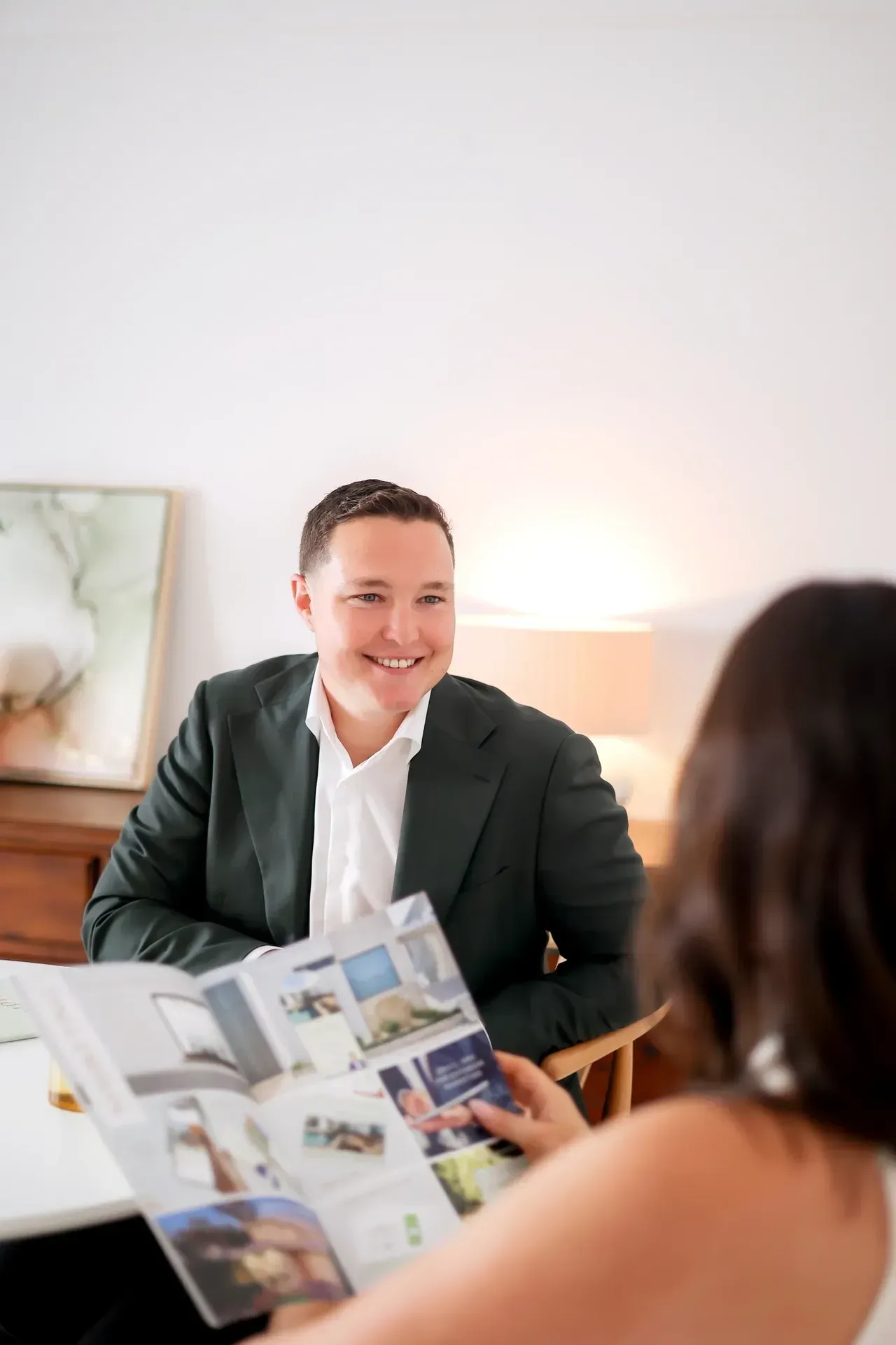 Man in blazer smiles, looking at a person holding a brochure. Light, indoor setting with artwork.