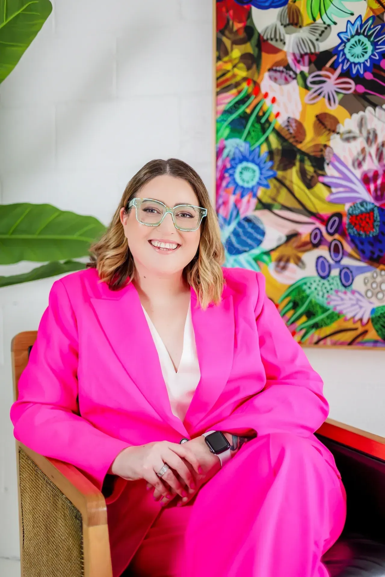 Woman in pink suit and glasses seated in front of colorful artwork.