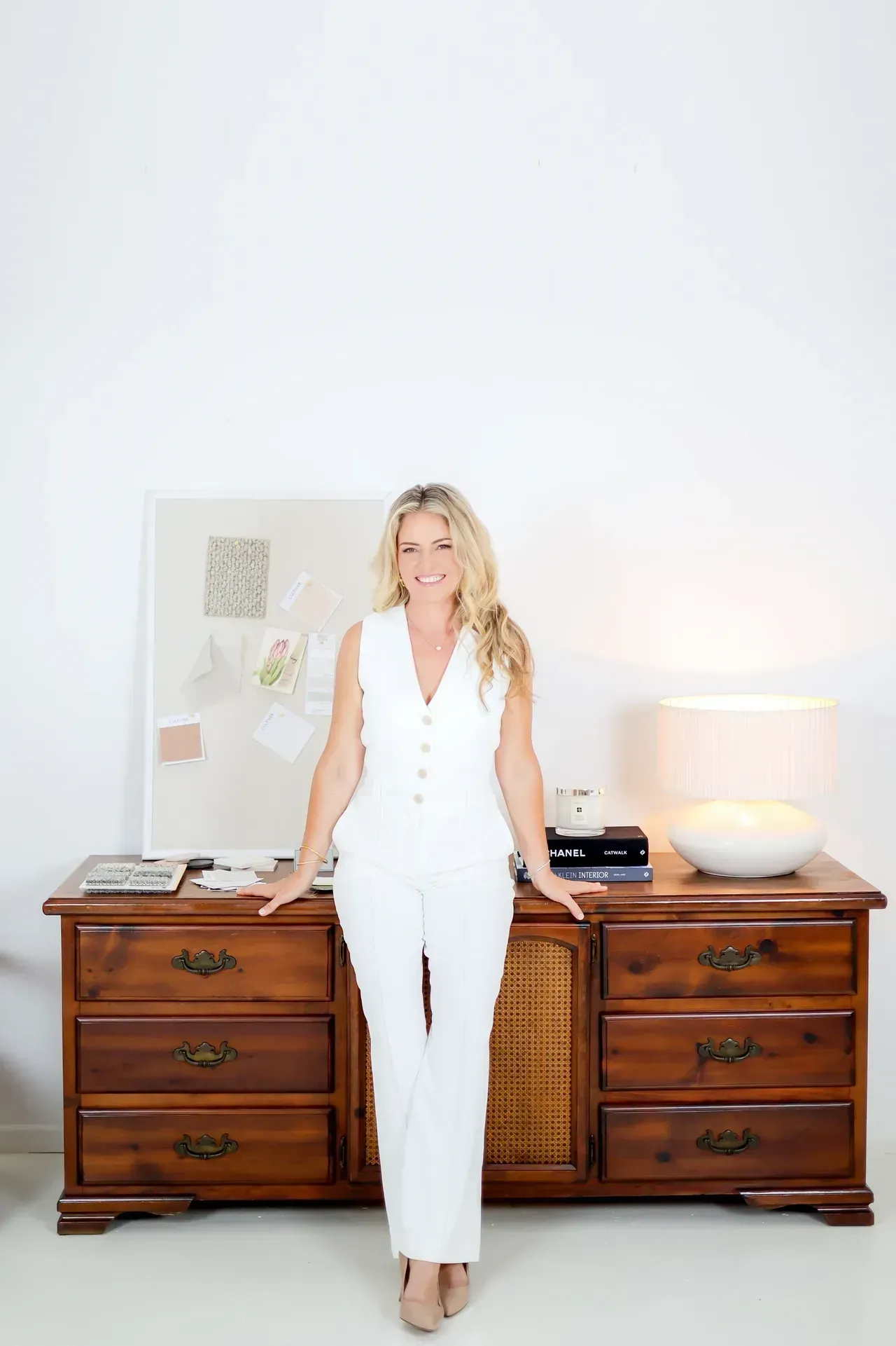 Woman in white suit leans on a wooden dresser. Bright, white background.