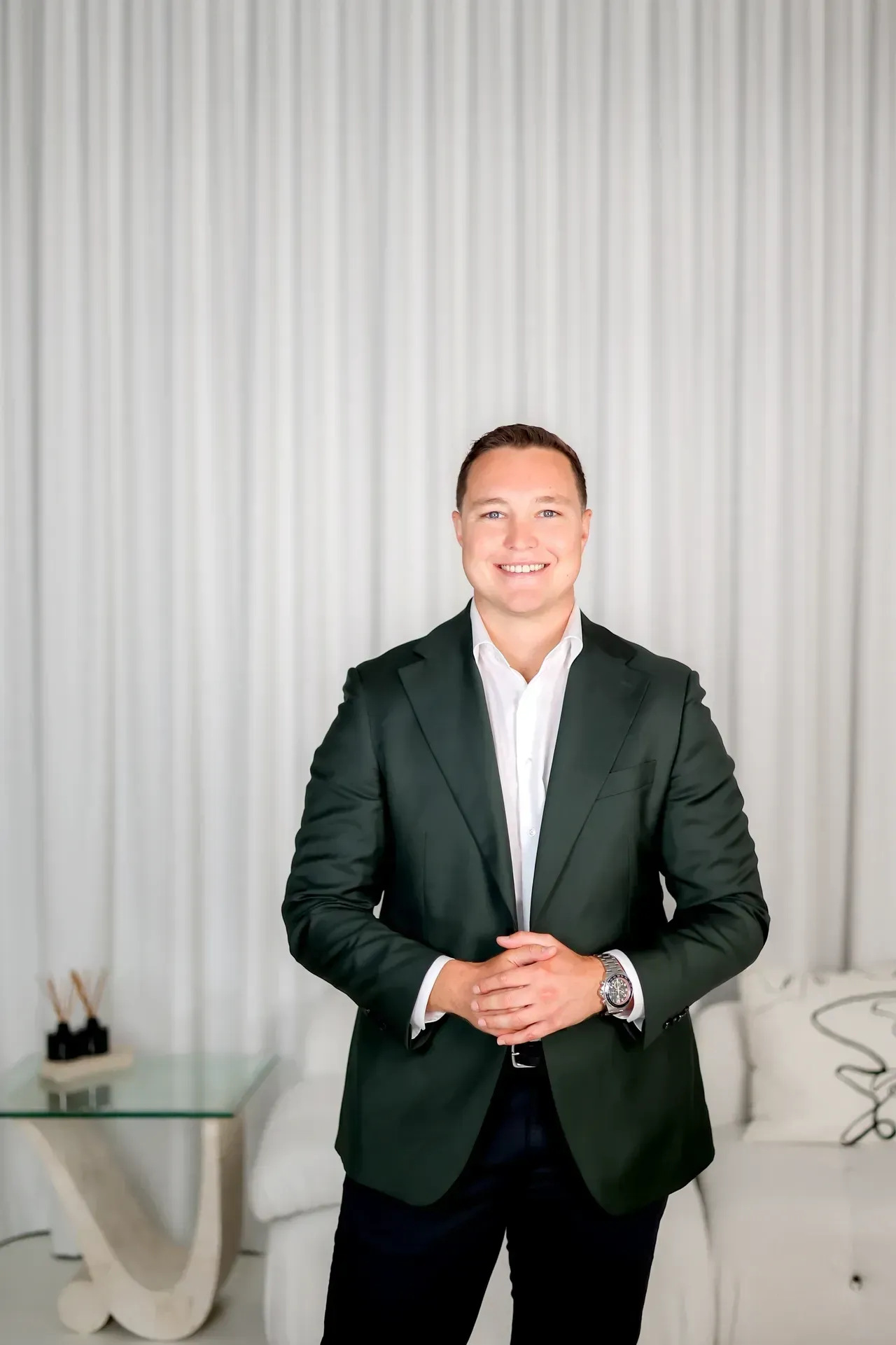 Man in green blazer smiles, arms crossed, in a white-walled room.
