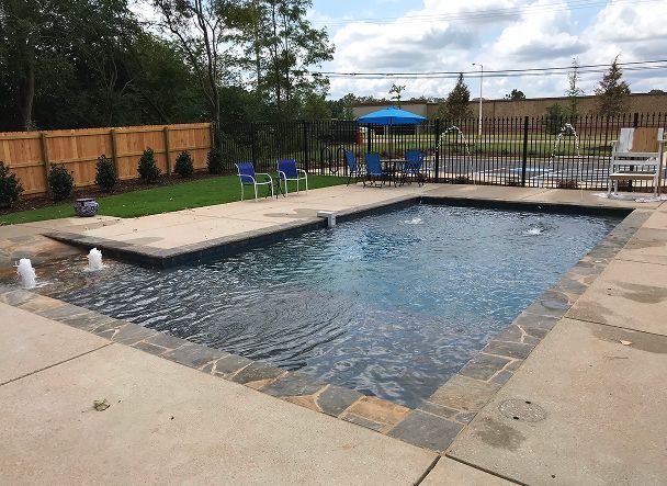 Swimming pool with water fountains, surrounded by concrete and stone. Chairs and fence in the background.