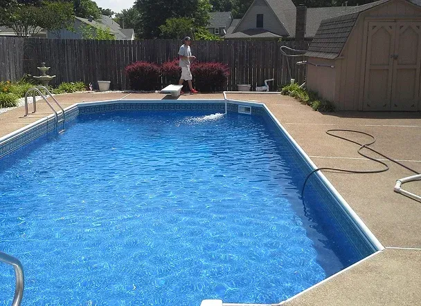 Rectangular pool with blue water. A person stands nearby. Concrete patio, wooden fence.