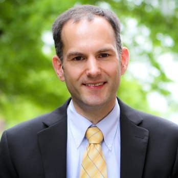 Man in a suit and tie smiling outdoors in front of a green background.