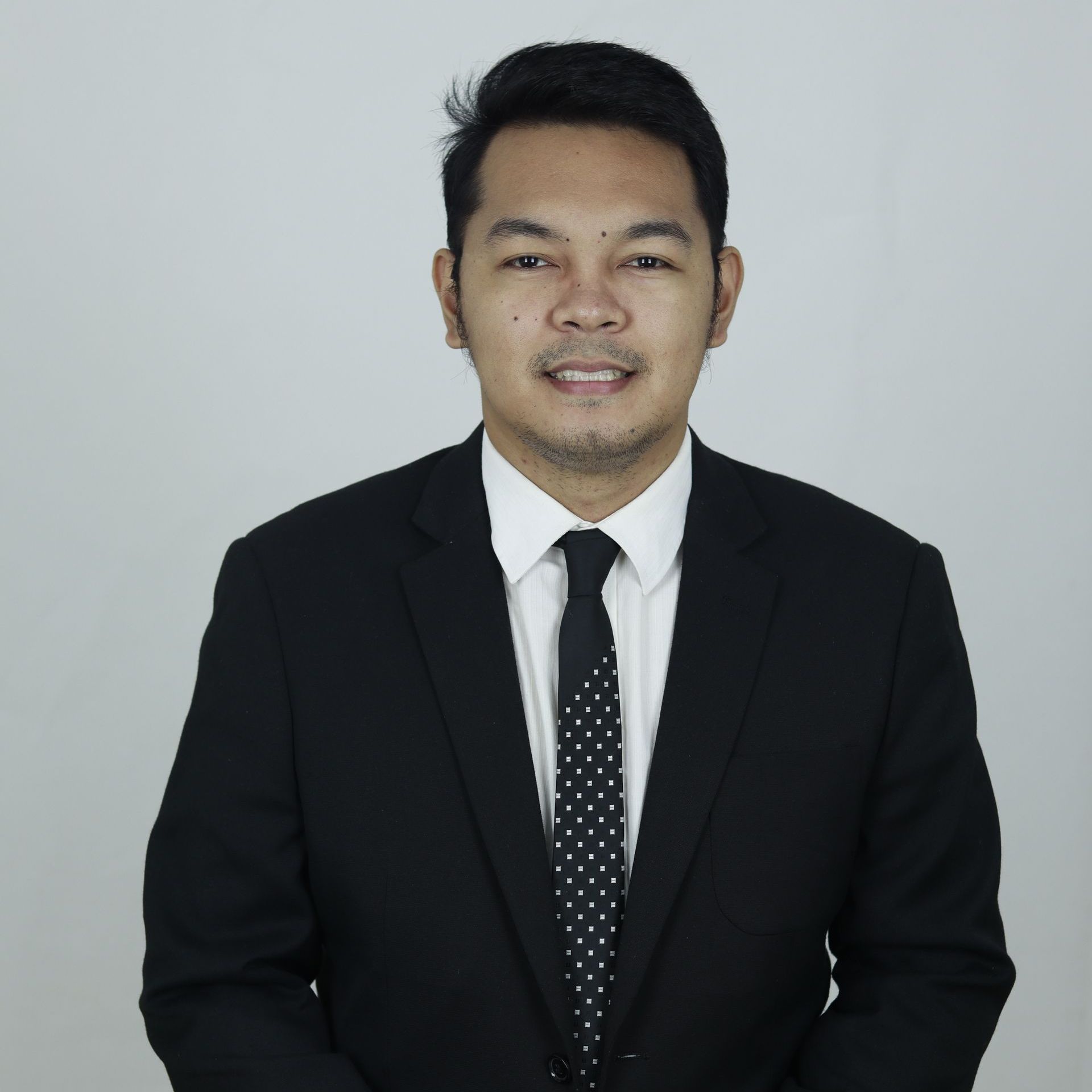 Man in a black suit and tie, smiling at the camera. White backdrop.