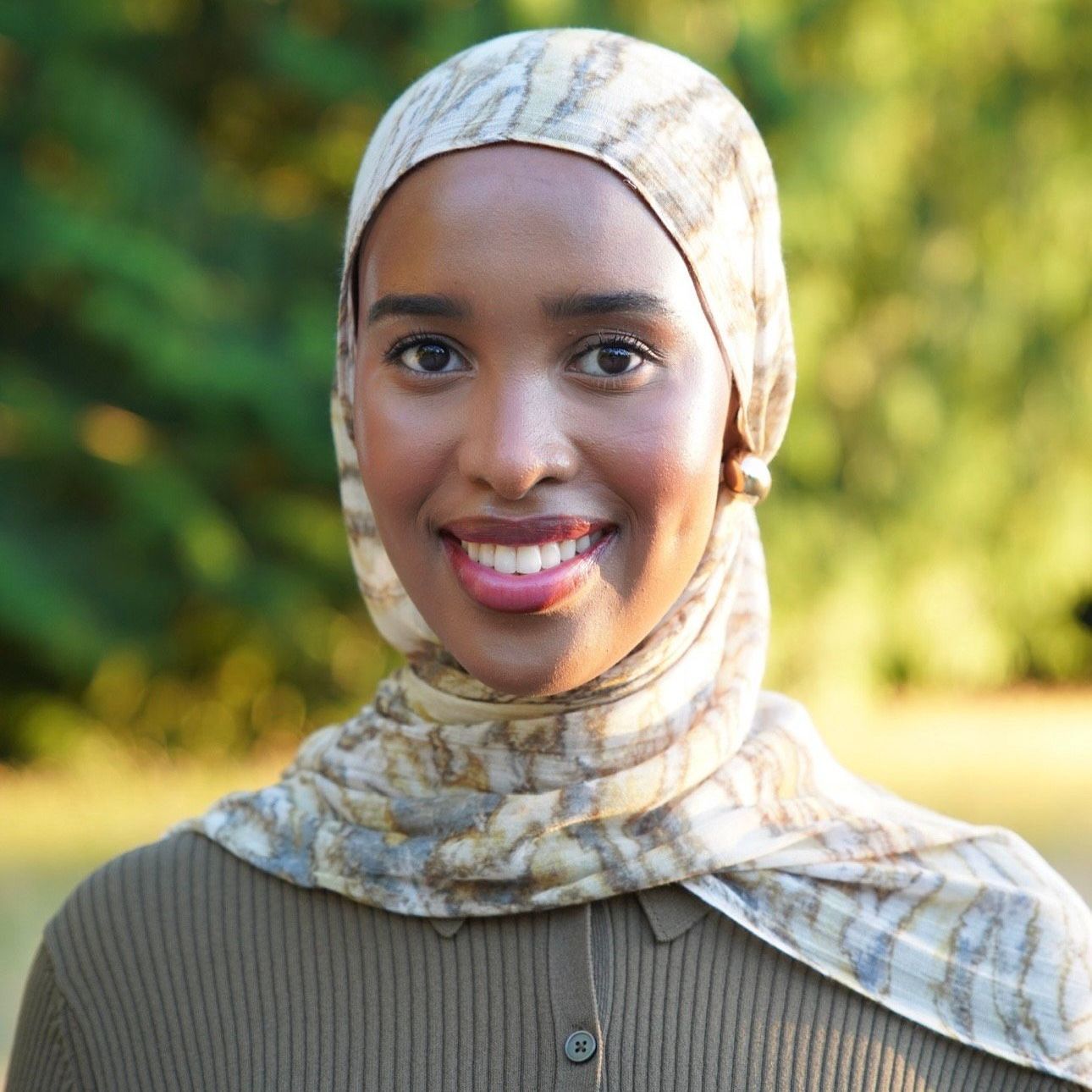 Woman with a hijab smiling outdoors, wearing an olive sweater.