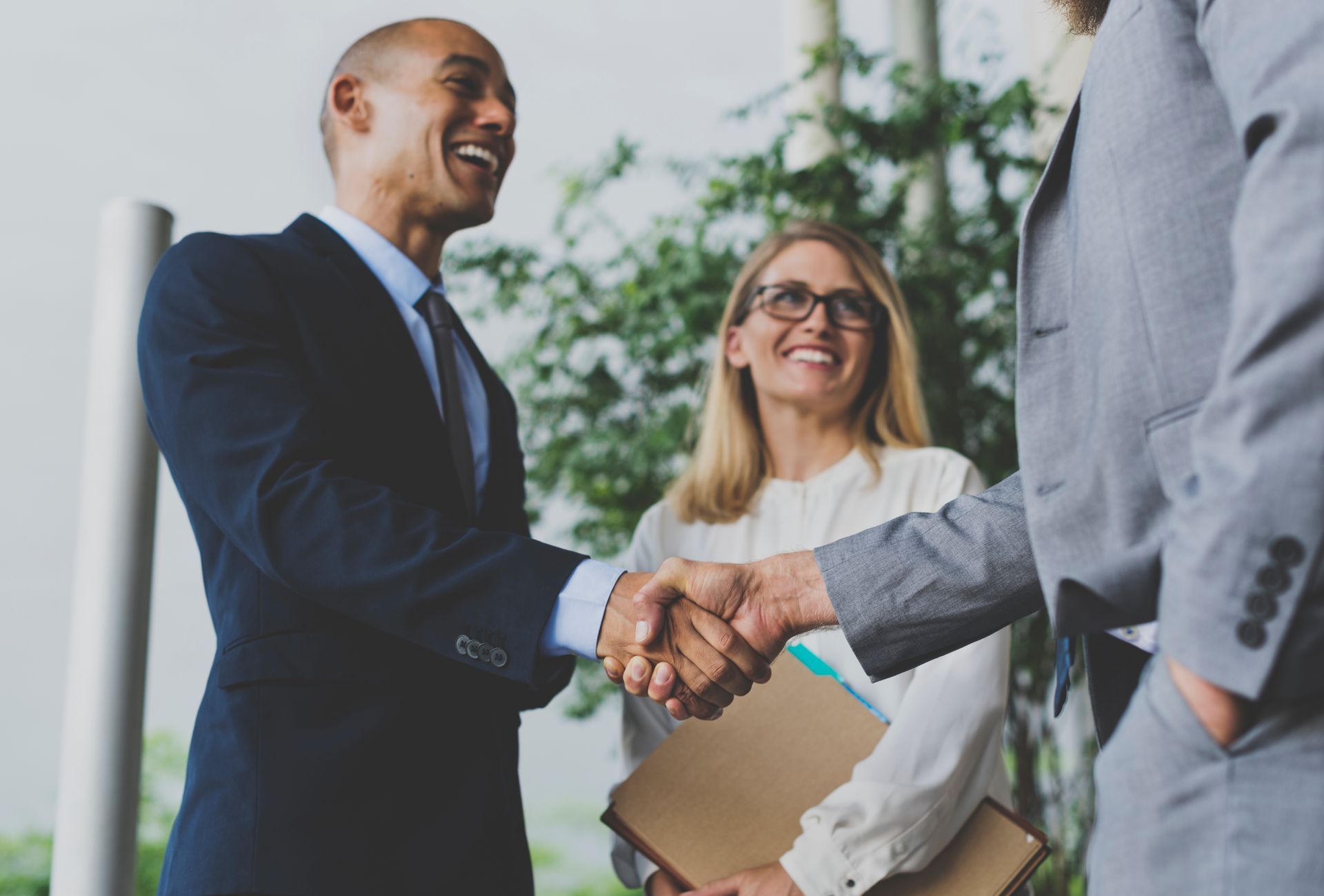 Three business people in suits shake hands, smiling, in an office setting.