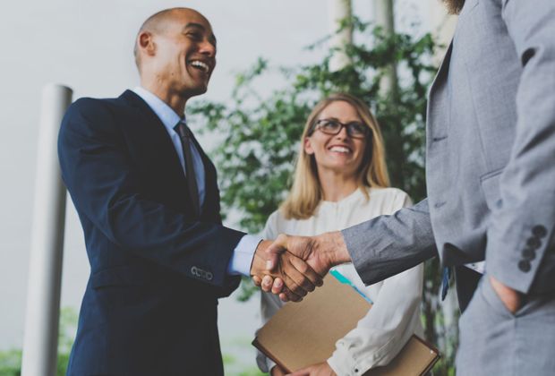 Three business people in suits shake hands, smiling, in an office setting.