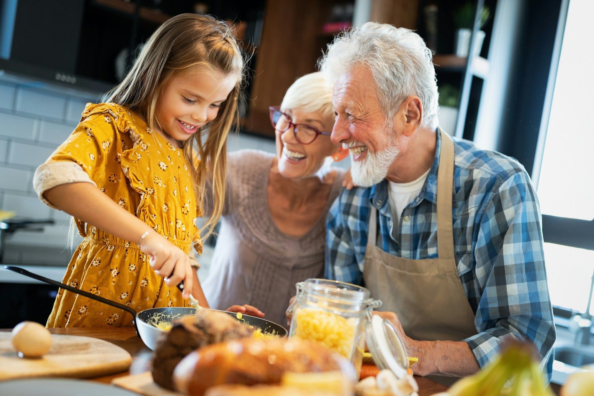 Girl and grandparents cooking together; girl stirs food in pan, all smiling in kitchen.