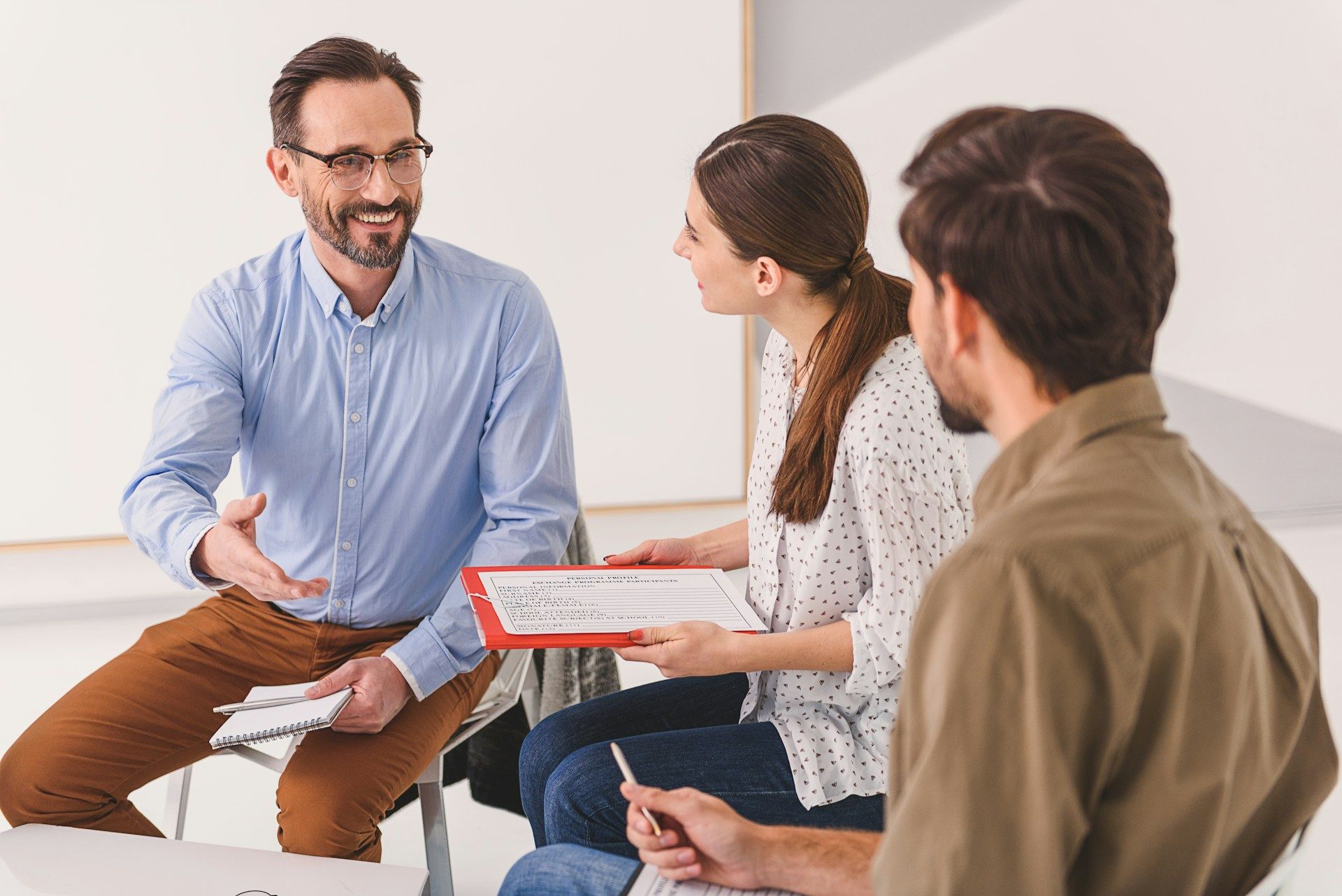 Group therapy session; a therapist and two clients in a bright room, discussing papers, smiling.