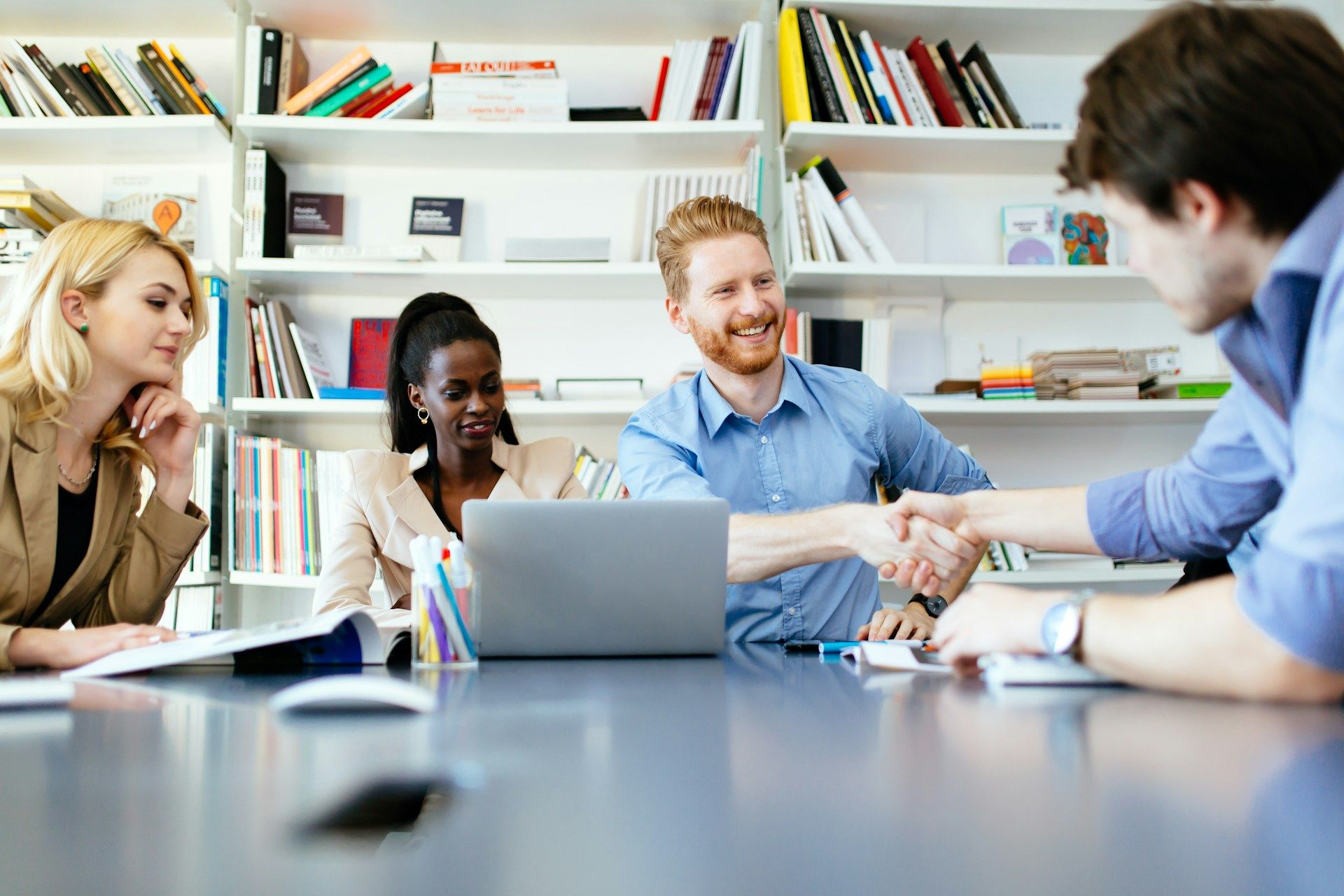 Four people in an office at a table, one shakes hands with another, smiles, books in background.