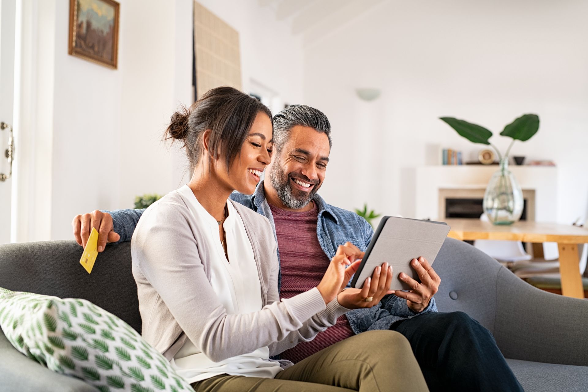 Couple on sofa looking at tablet, woman pointing, man smiling. Living room setting.