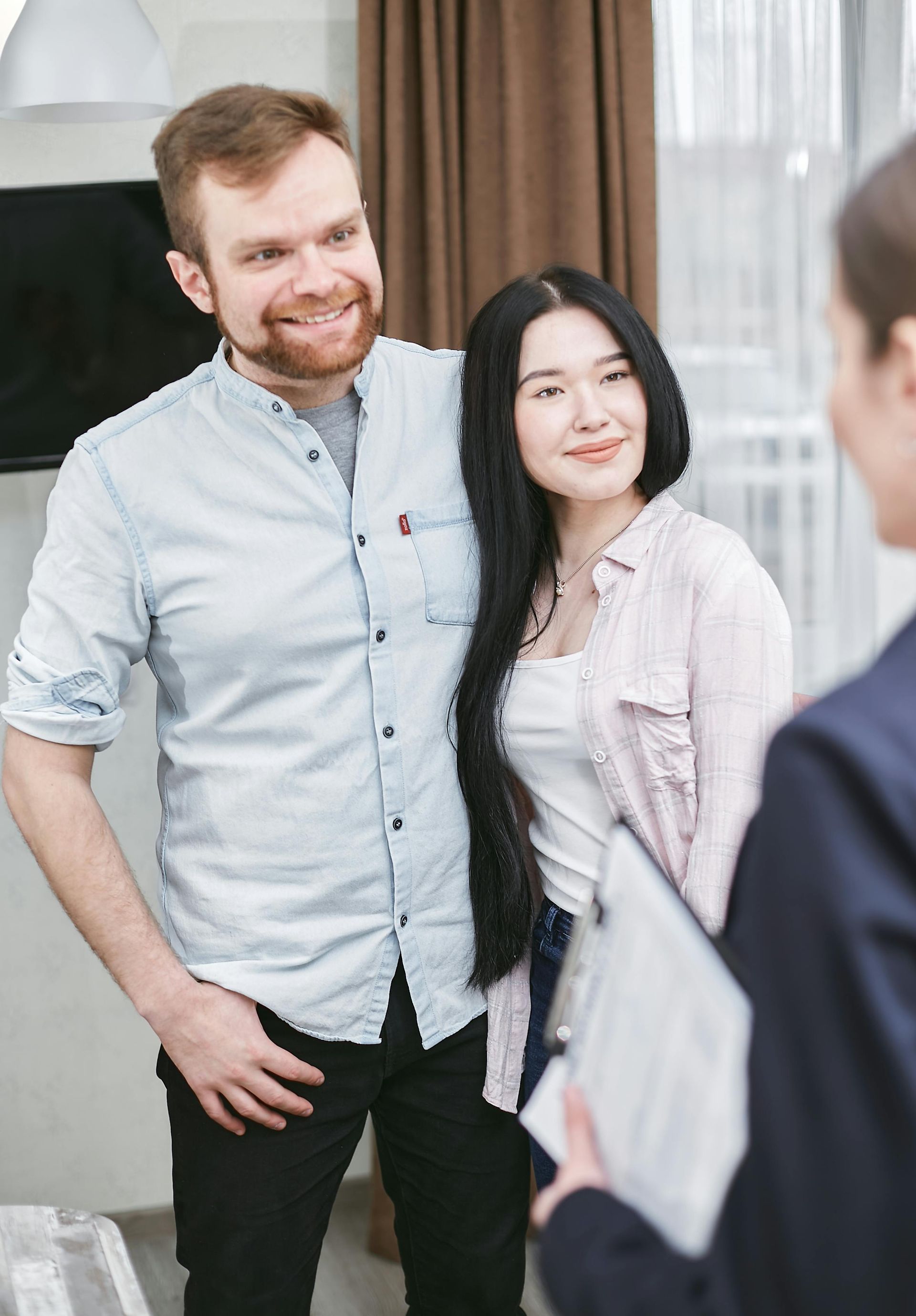 Couple smiles with real estate agent in a home, brown curtains, and TV.