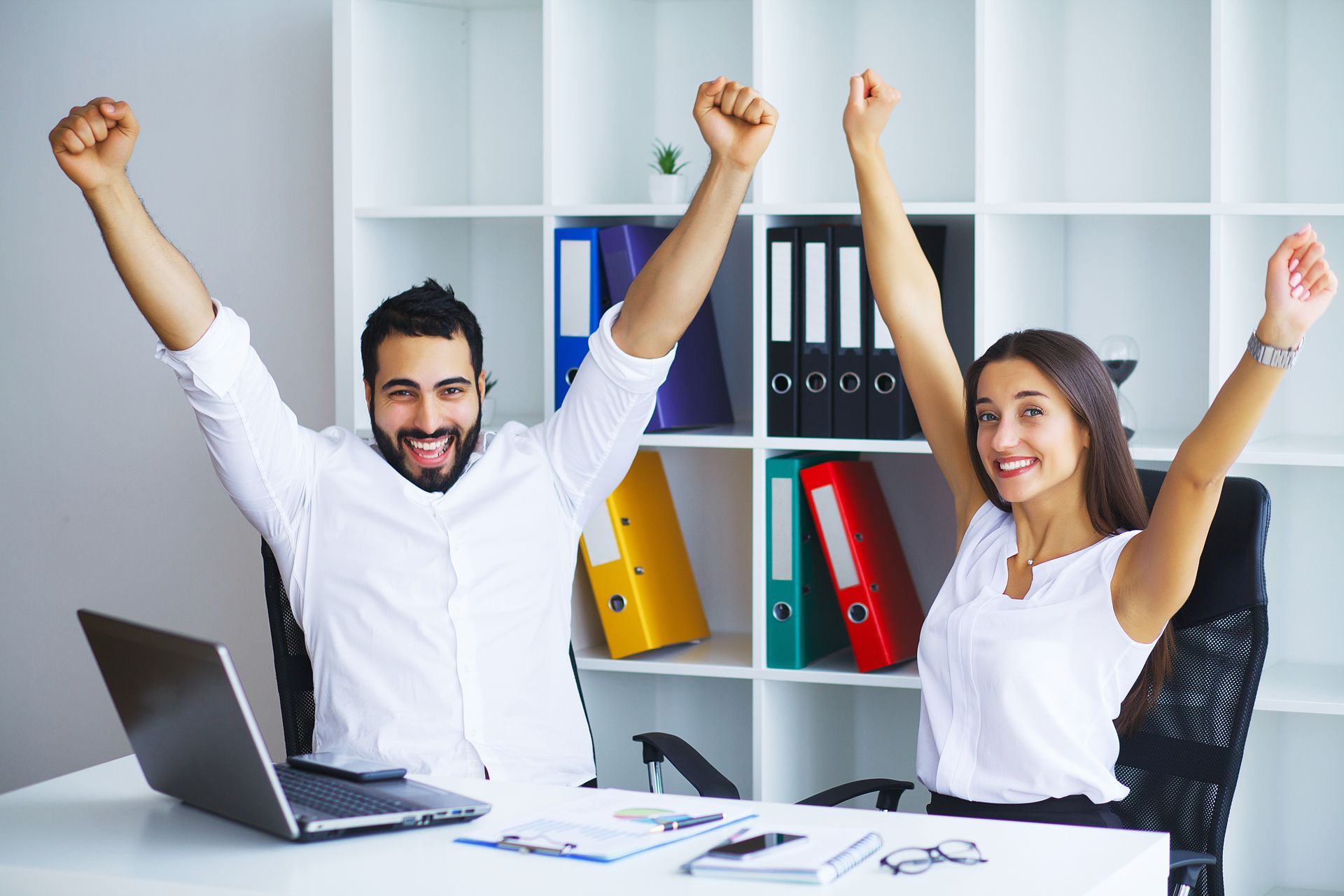 Man and woman in office raise arms in celebration; laptop and file folders in background.