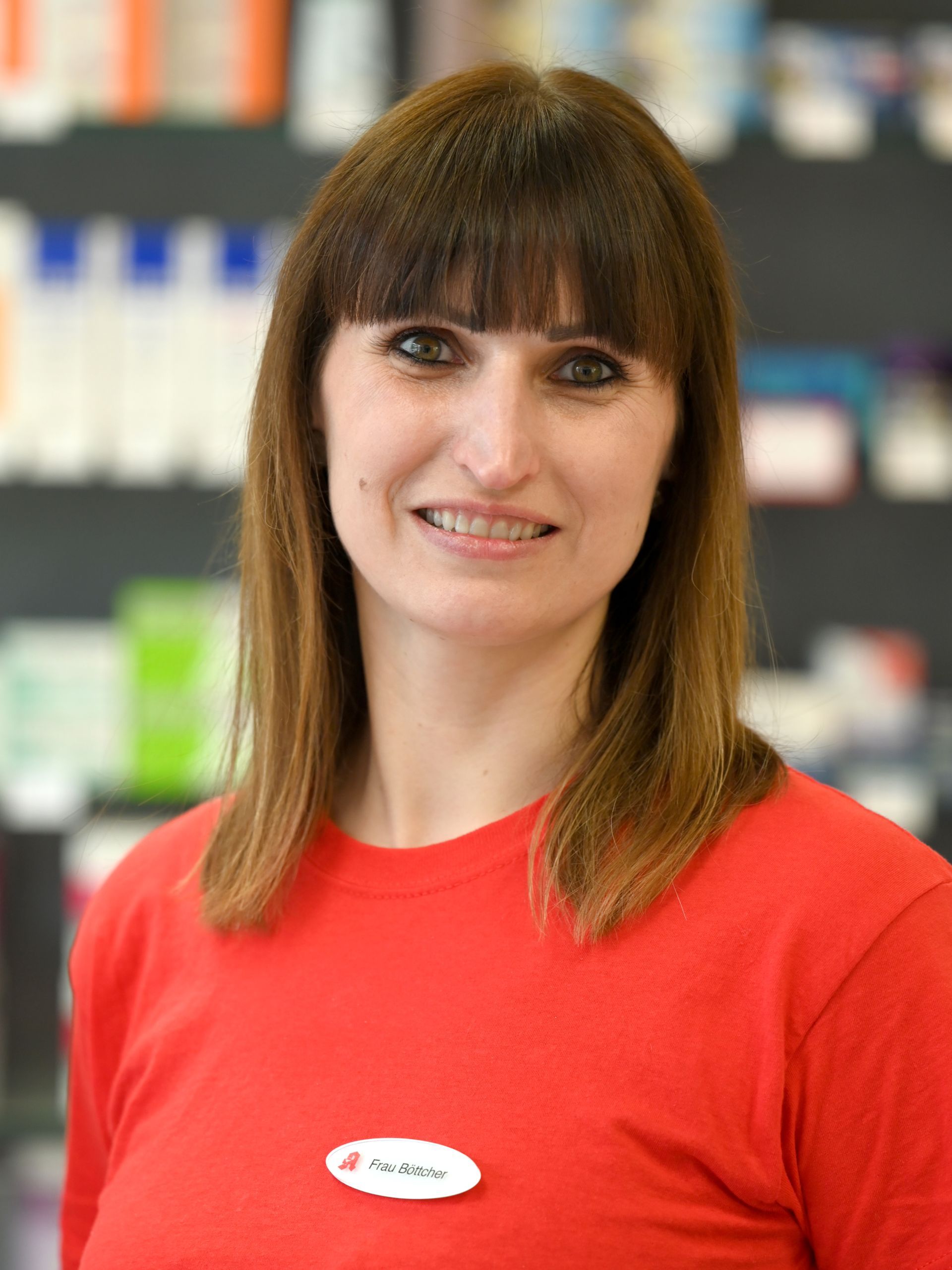 Frau im roten Hemd lächelt in einer Apotheke, Namensschild sichtbar, Regale im Hintergrund.
