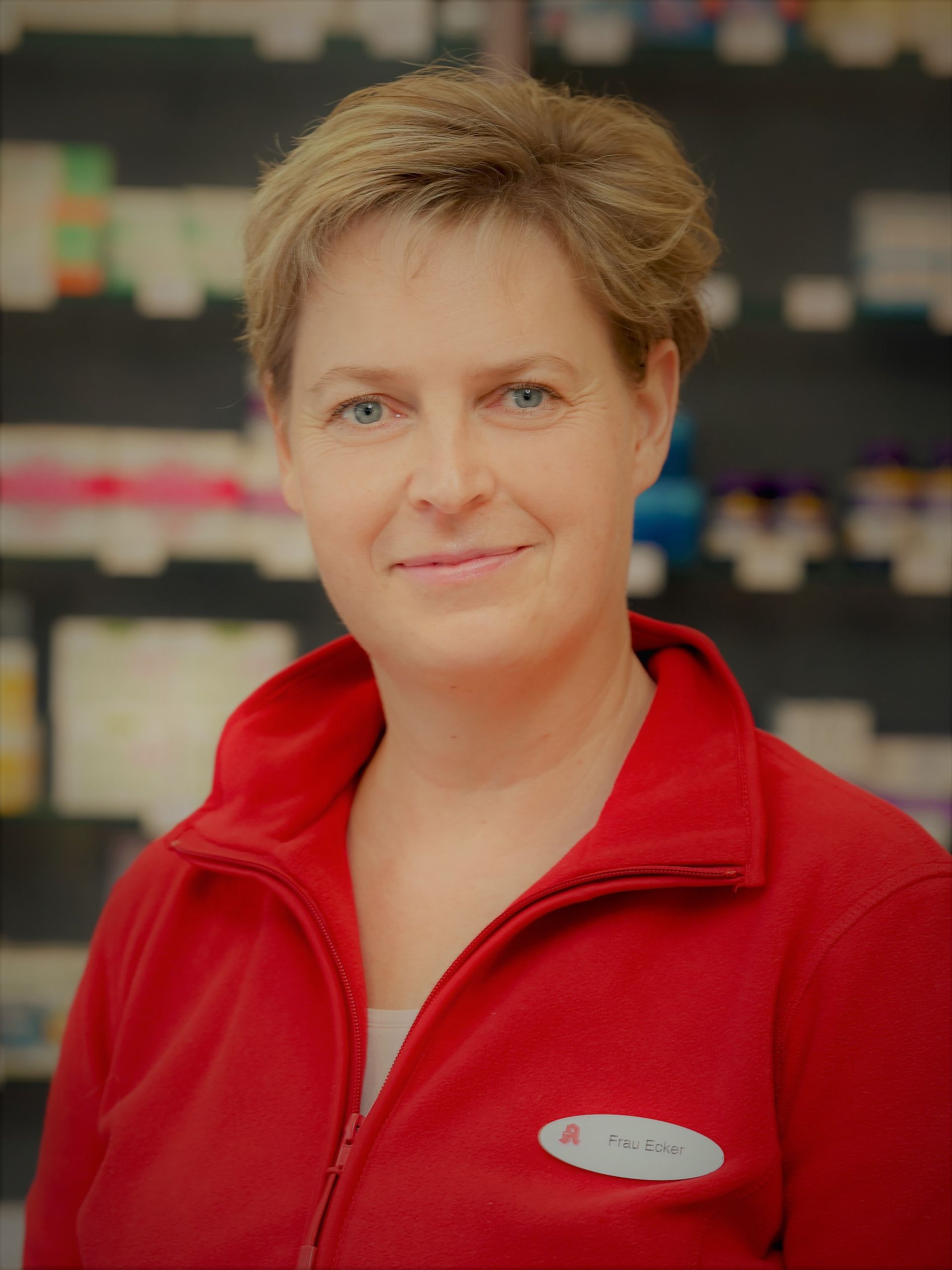 Frau mit kurzen blonden Haaren, trägt eine rote Jacke und lächelt in einem Laden.