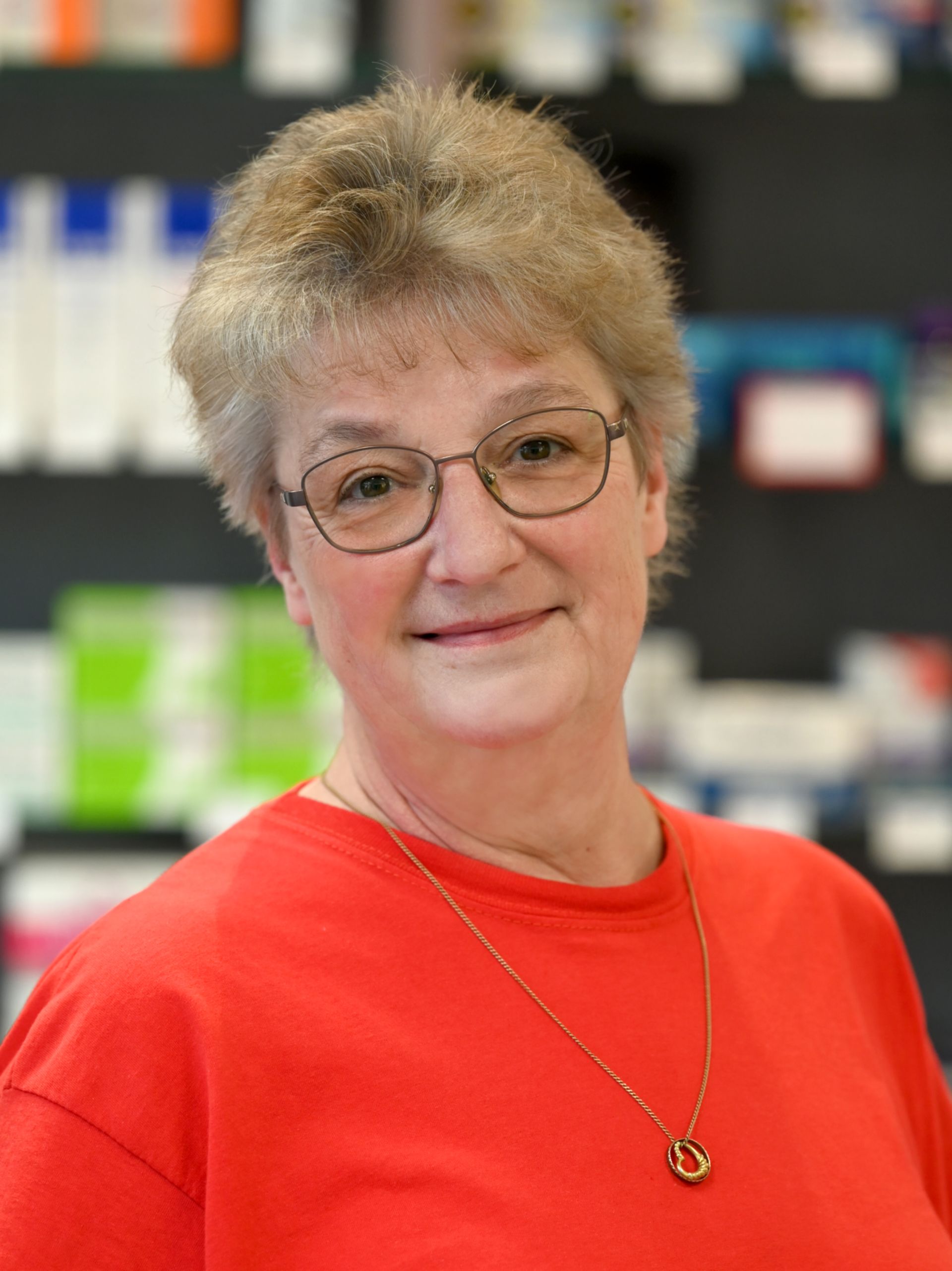 Frau mit Brille und rotem Hemd in einer Apotheke lächelt in die Kamera.