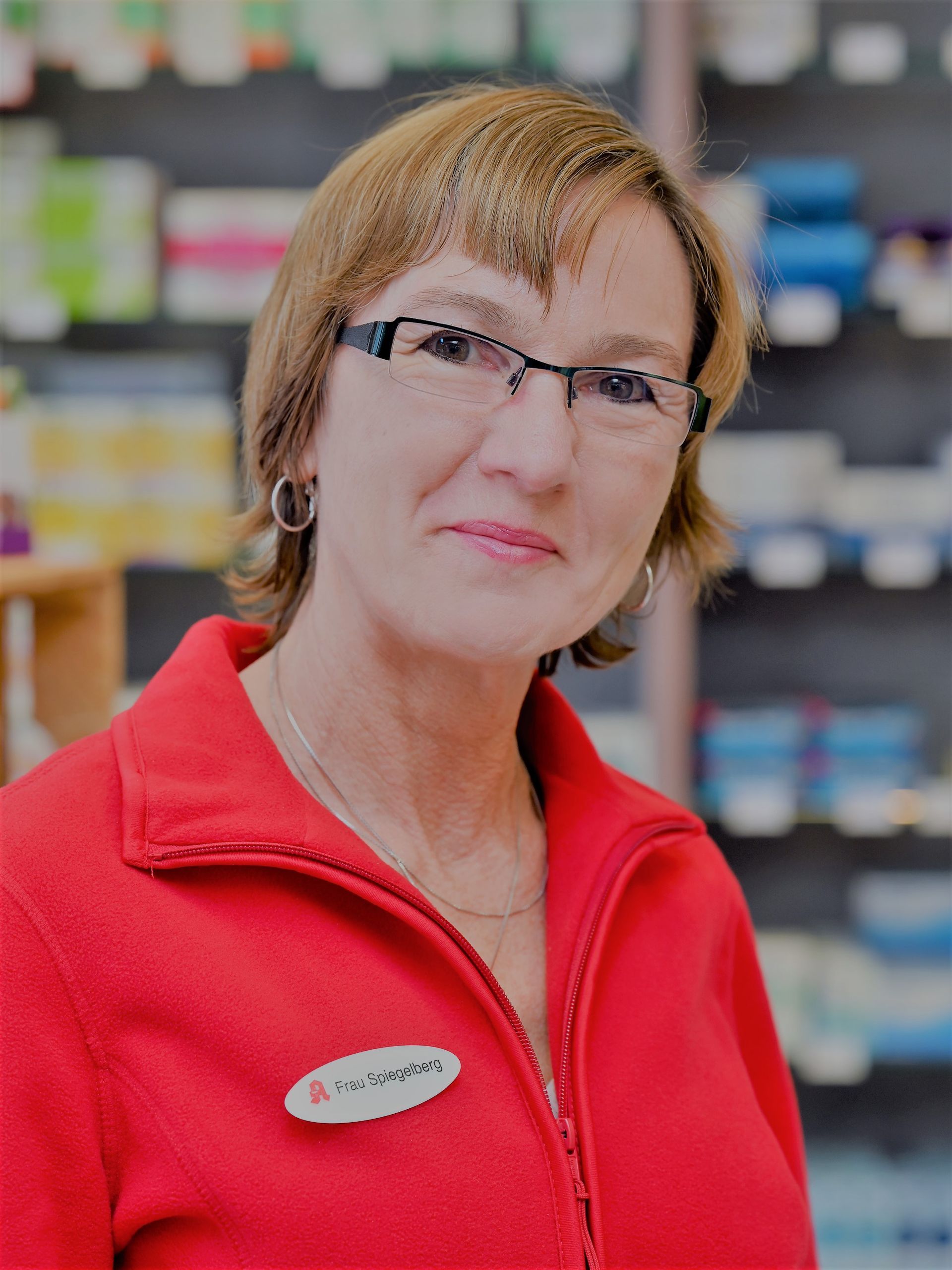 Frau in rotem Hemd und Brille lächelt in einer Apotheke und trägt ein Namensschild.