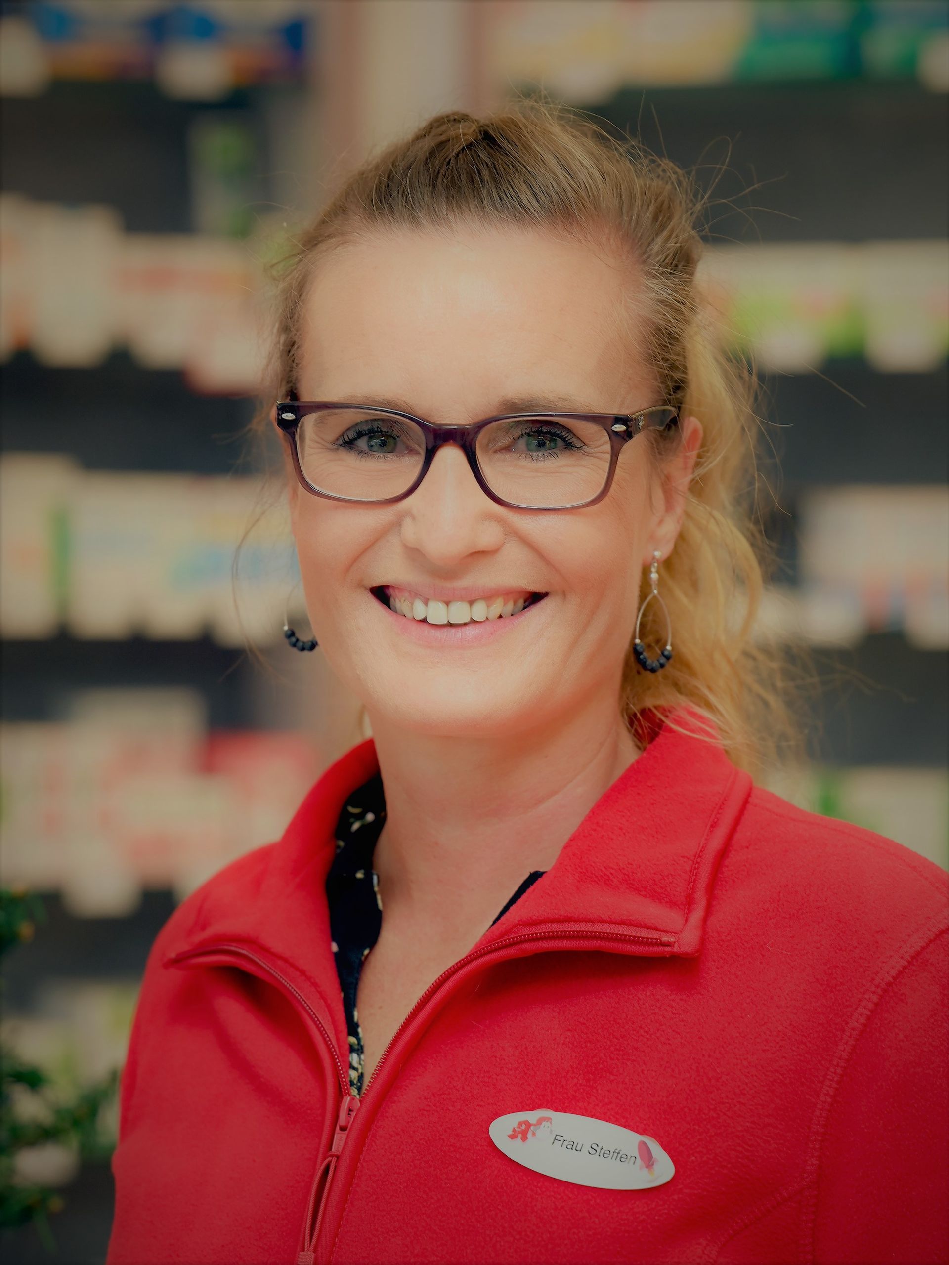 Frau in rotem Hemd, Brille, lächelnd, in einer Apotheke.