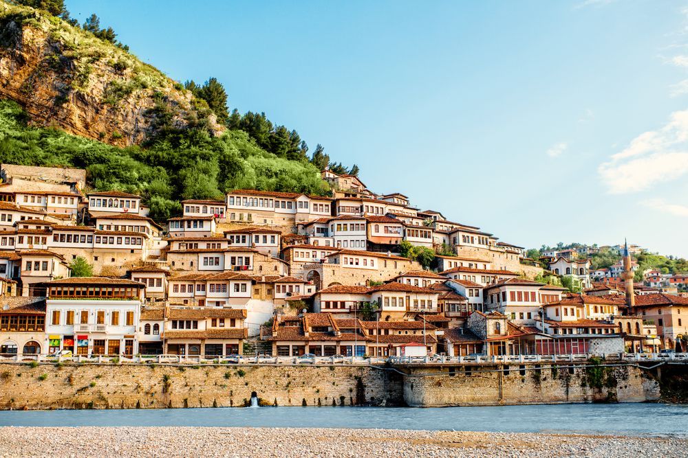 Whitewashed buildings with red-tiled roofs cascade down a hillside. Green trees and a blue sky provide a backdrop.