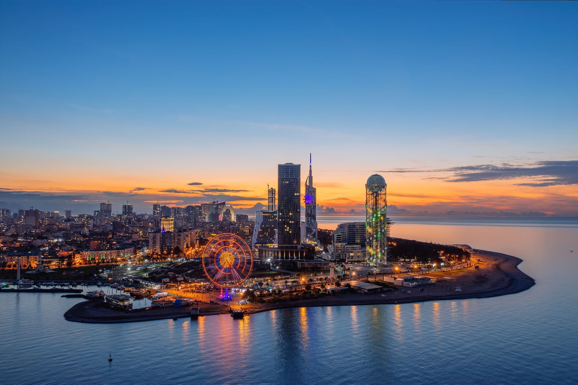City skyline at dusk, lights reflecting on calm water. Ferris wheel and modern buildings on a peninsula.