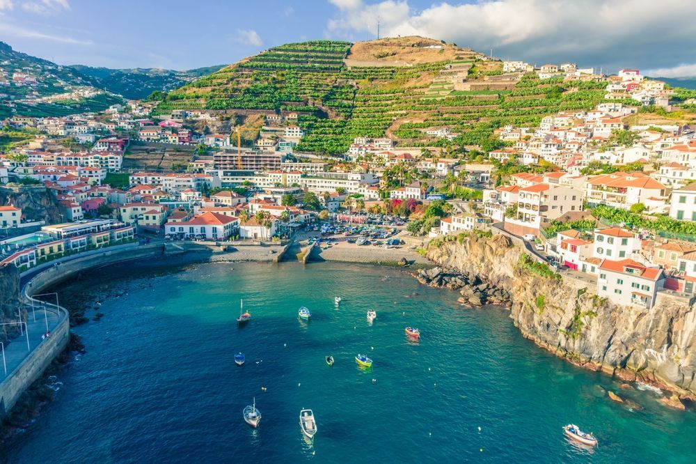 Coastal town nestled on hillside, blue water, boats, white buildings with red roofs, lush green terraces.