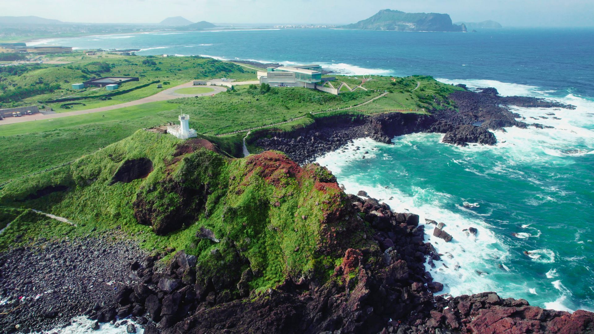 Coastal view of a green, rocky cliff with a white structure, meeting turquoise water. Distant land visible.